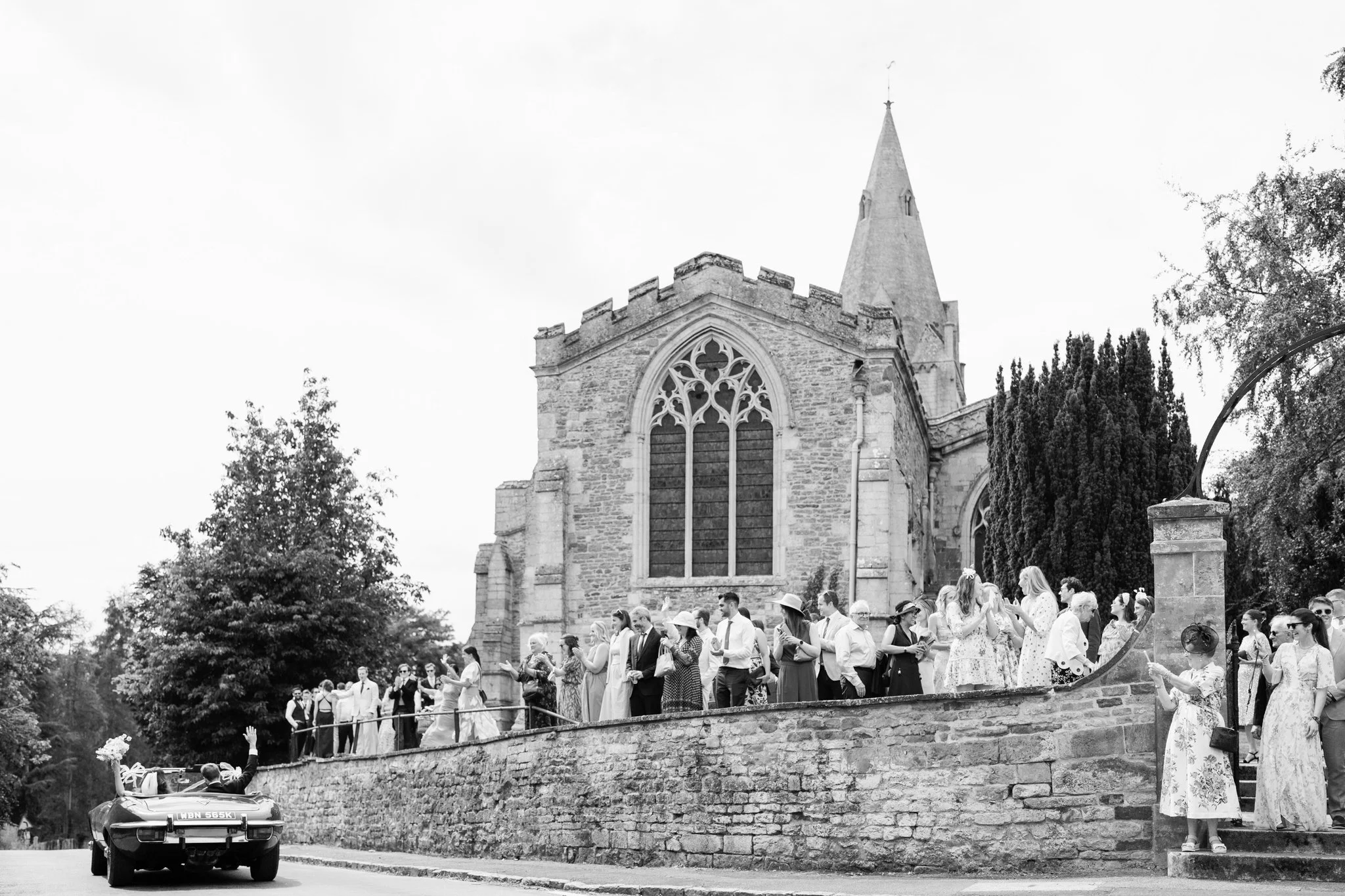 Bride and groom leave church in vintage car and wave at guests during english countryside wedding