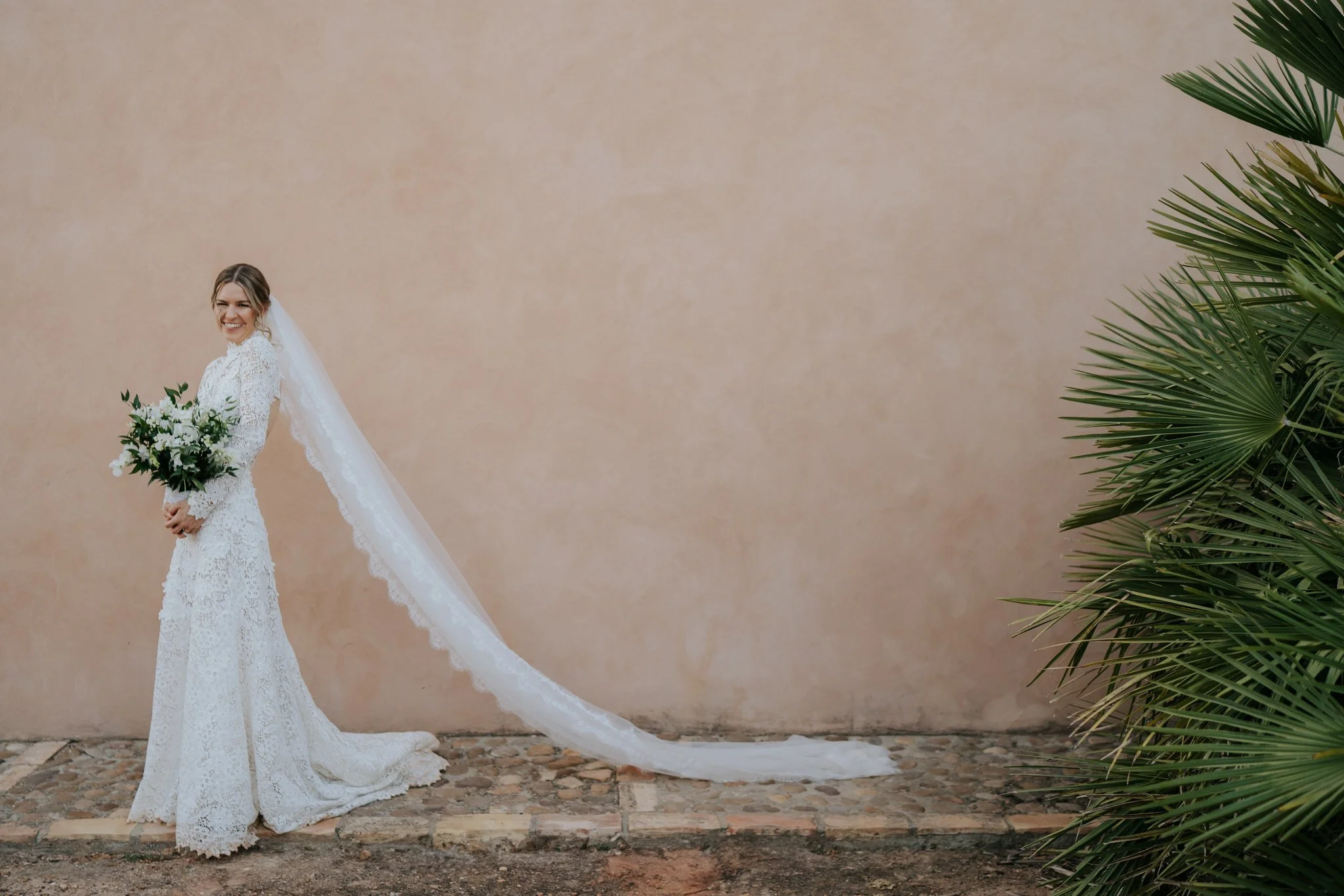 Bride poses elegantly in front of pink wall at outdoor wedding in Seville as her veil runs the length of the frame