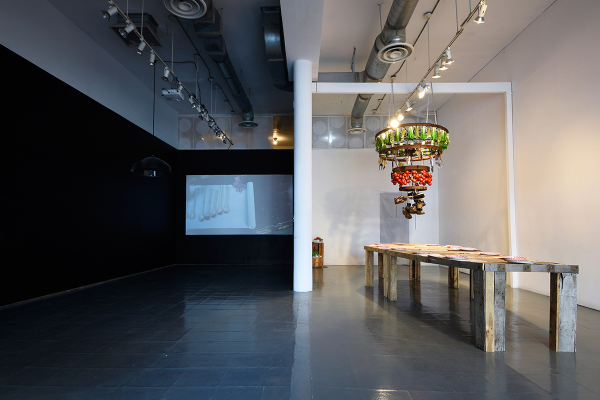 The Produce Party (2017) , commissioned by Artcite Inc, was shown in Walks of Survivance , a two-person exhibition curated by Srimoyee Mitra. In this installation, the ambience of a locally sourced soirée is breached by a series of interventions. A coffin-shaped dining table, made from fruit-crates, pays homage to migrant farmworkers in Canada. Thirteen disposable paper plates are nailed to its surface. Overhead, a chandelier adorned with soiled shoes, gloves and plastic produce dangles with the remnants of labour and loss. Photos by Joshua Babcock, Programming Coordinator, Artcite Inc. 
