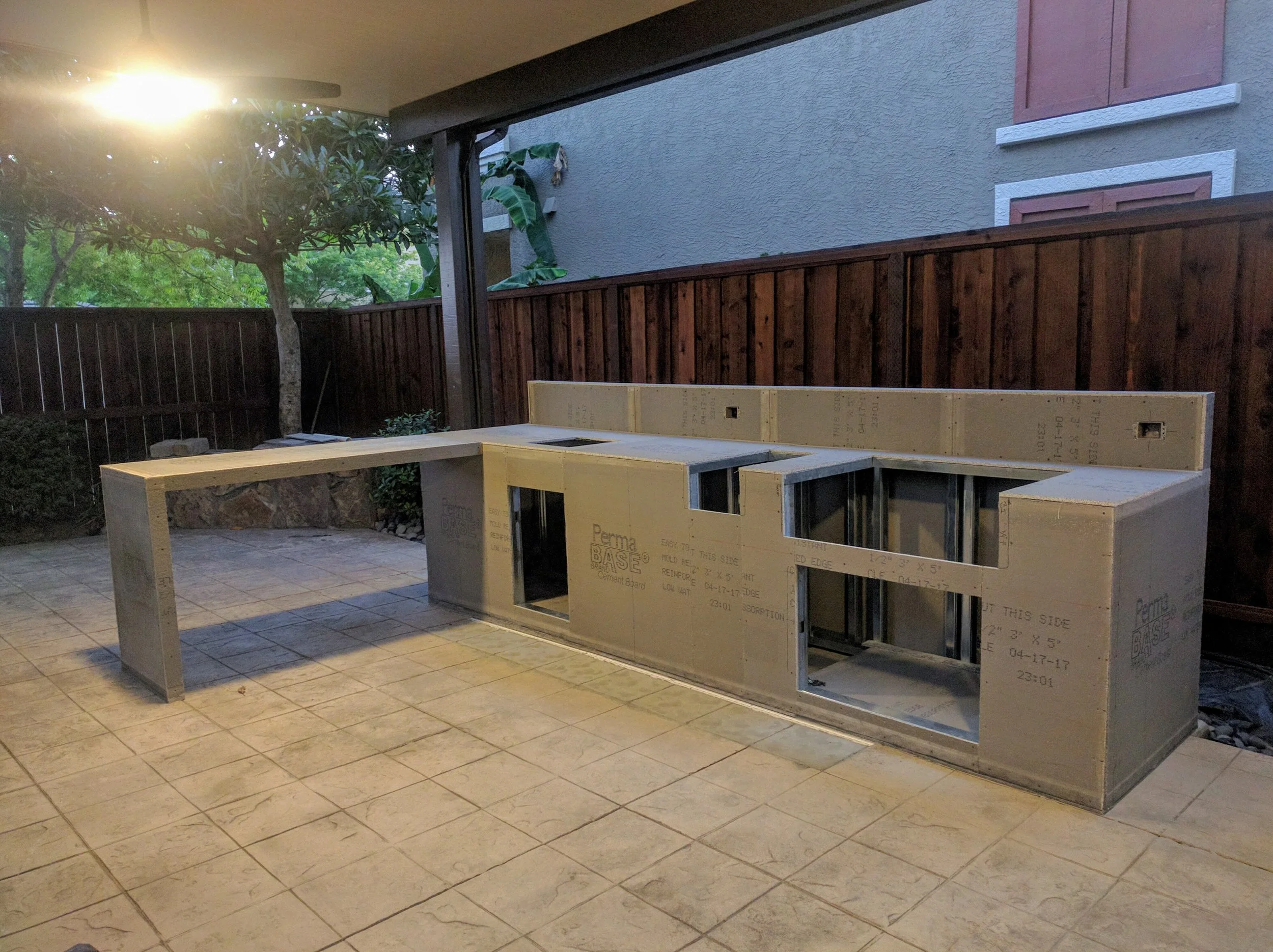 Construction of an outdoor kitchen with framed cabinetry on a tiled patio, with a wooden fence, trees, and house wall in the background.