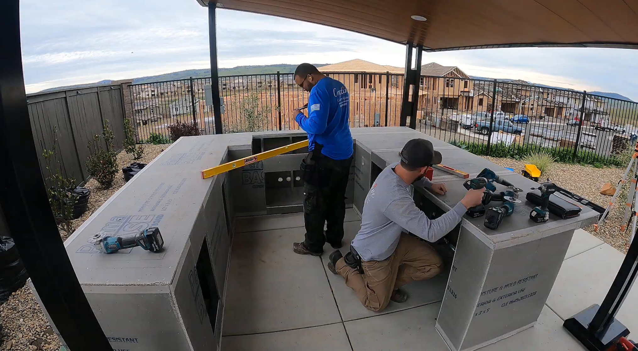 Two construction workers installing an outdoor kitchen counter, with tools and drill on the counter, and a yellow level tool placed across the counter.