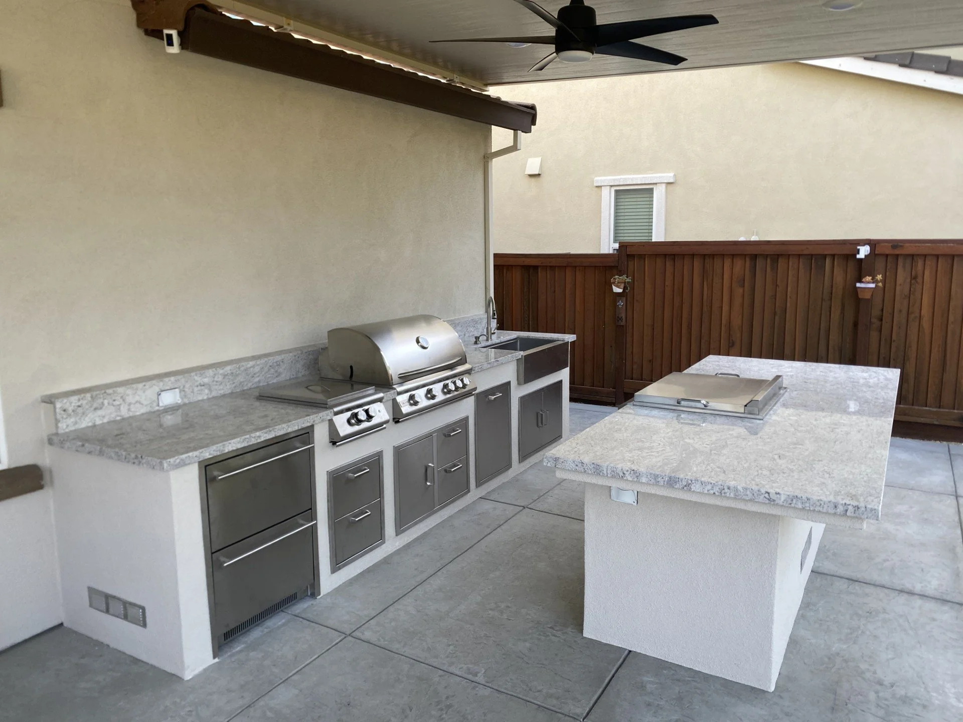 Outdoor kitchen with a granite counter, a stainless steel gas grill, a small sink, and a wooden fence in the background at a patio.