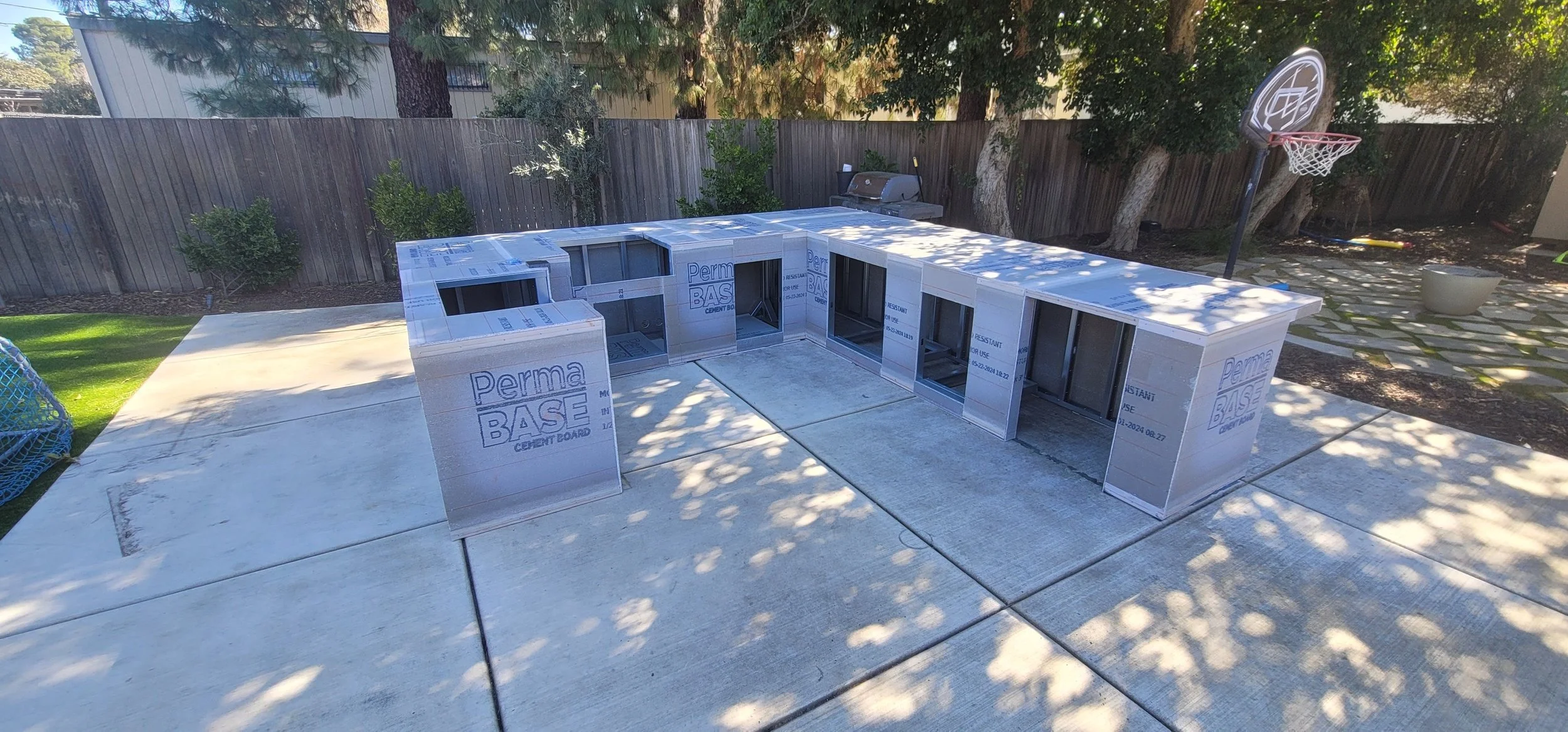 Construction of a small building using cement board on a backyard patio, with a basketball hoop and trees in the background.