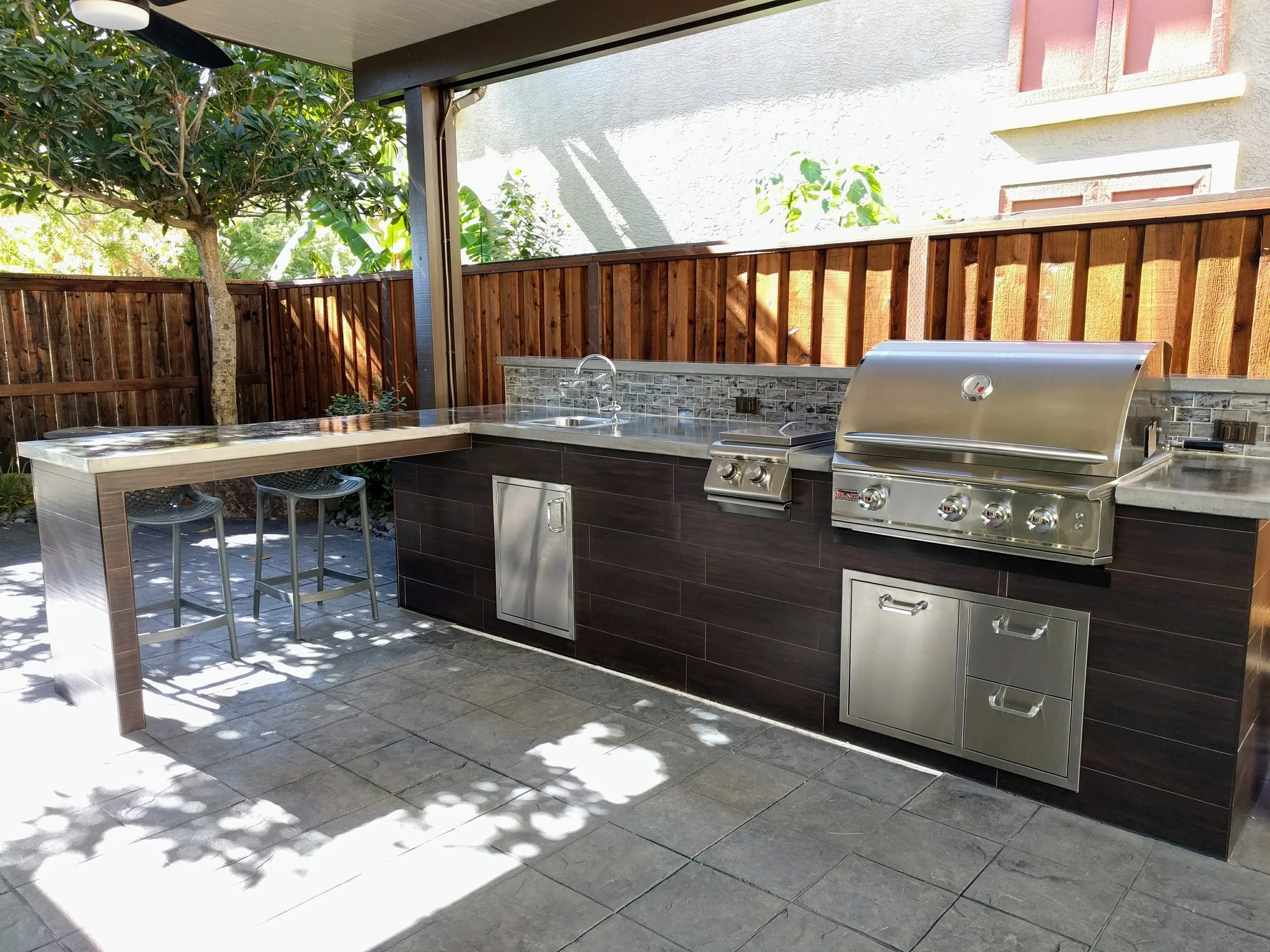 Outdoor kitchen with a stainless steel grill, sink, countertop, and bar seating, enclosed by a wooden fence with trees in the background.