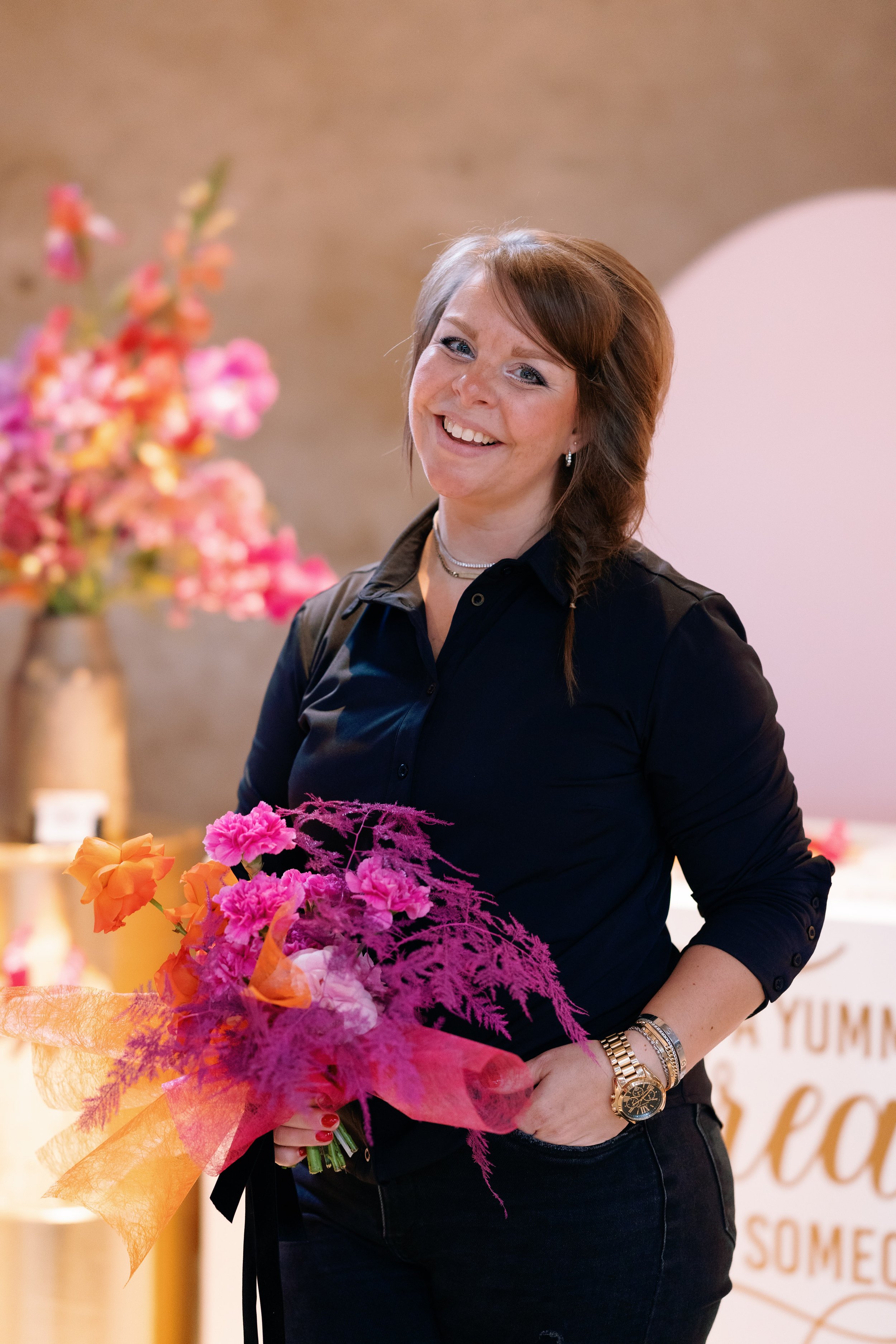 Vrouw met een kleurrijk boeket bloemen in haar hand, gekleed in donkere kleding, met een achtergrond van bloemendecoraties.
