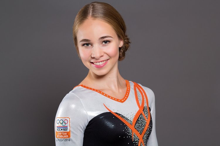 Portret van een jonge vrouw in een turnpak met oranje en zwarte accenten, glimlachend naar de camera. Op haar mouw staat een embleem met 'NOC*NSF'. Grijze achtergrond.