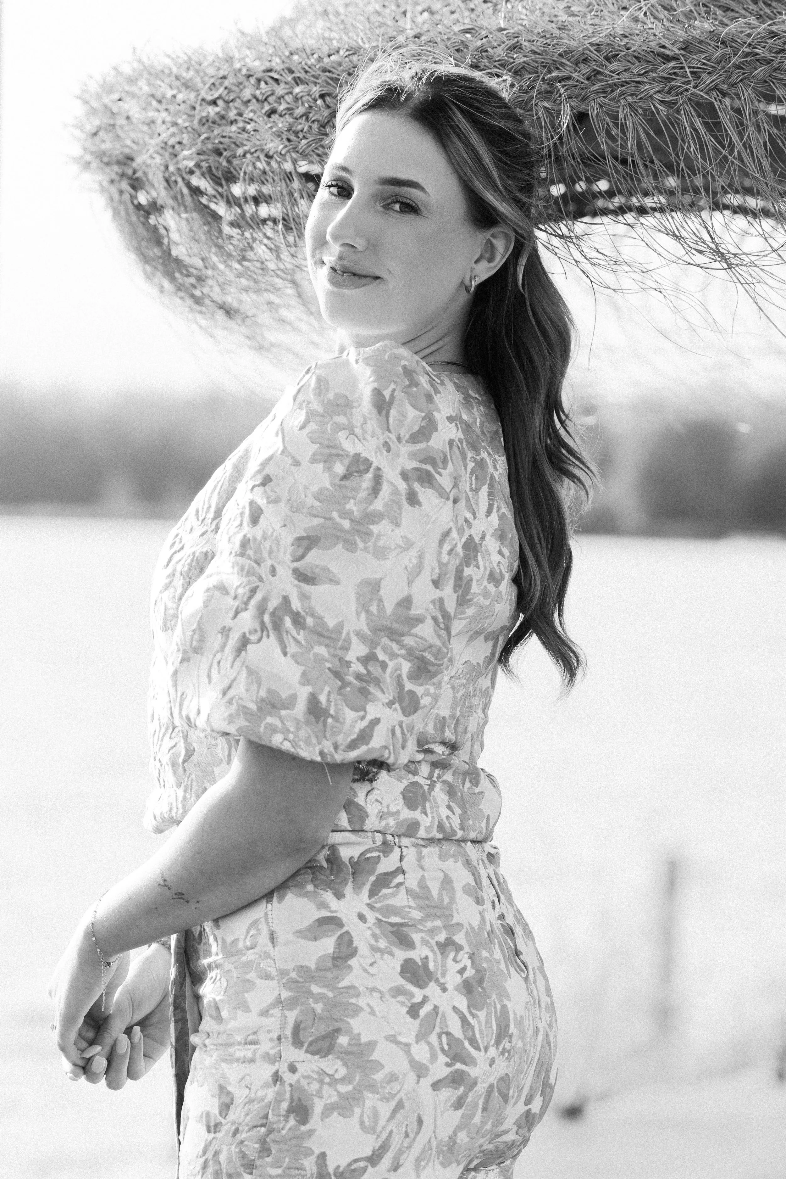 Zwart-wit foto van een vrouw met lang haar die naar de camera kijkt, buiten, met een strooien zonnehoed op haar schouder.