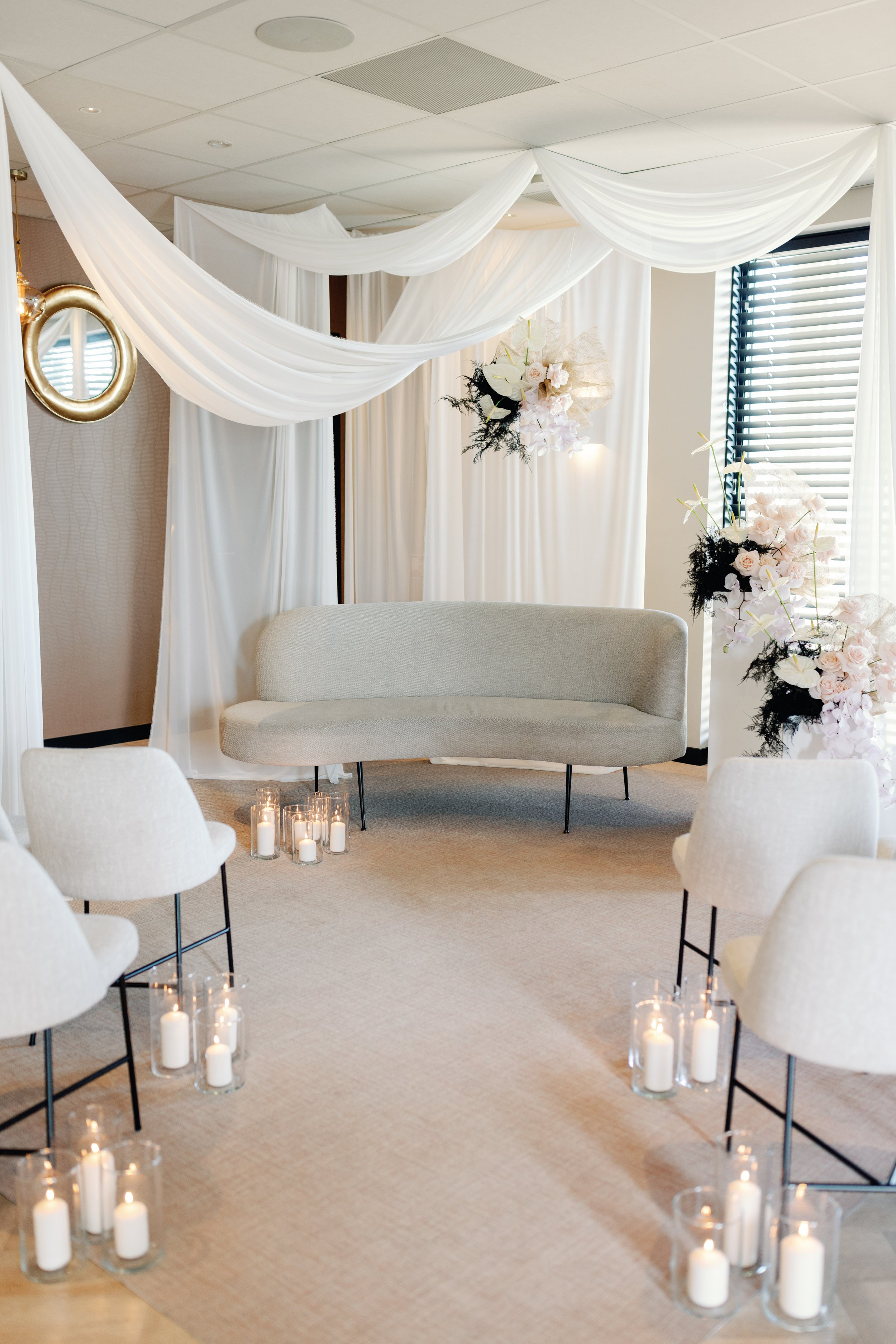 Een huwelijksceremonie ruimte met witte gordijnen, een moderne gebogen bank, witte stoelen, bloemenarrangementen en kaarsen in glazen houders op de vloer.