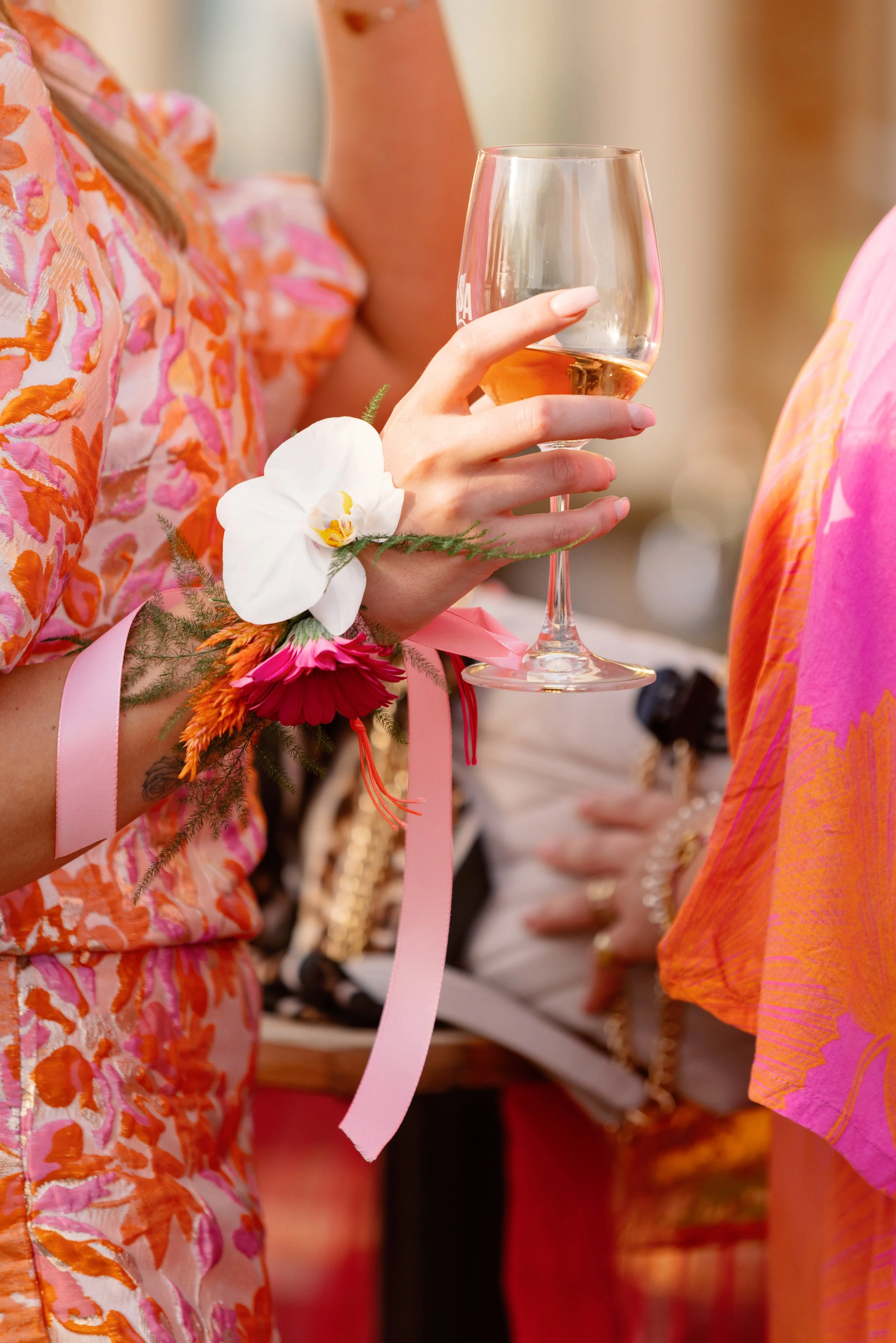 Close-up van een vrouw met een bloemenarmband die een glas rosé wijn vasthoudt tijdens een feest of bruiloft, met andere feestgangers op de achtergrond.