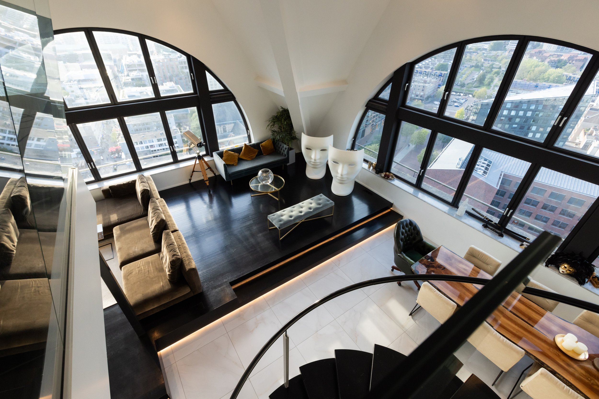 Luxe woonkamer met grote ramen, een bruine bank, twee grote sculptuurstoelen en een glazen salontafel. Er is een houten eethoek en een wenteltrap.
