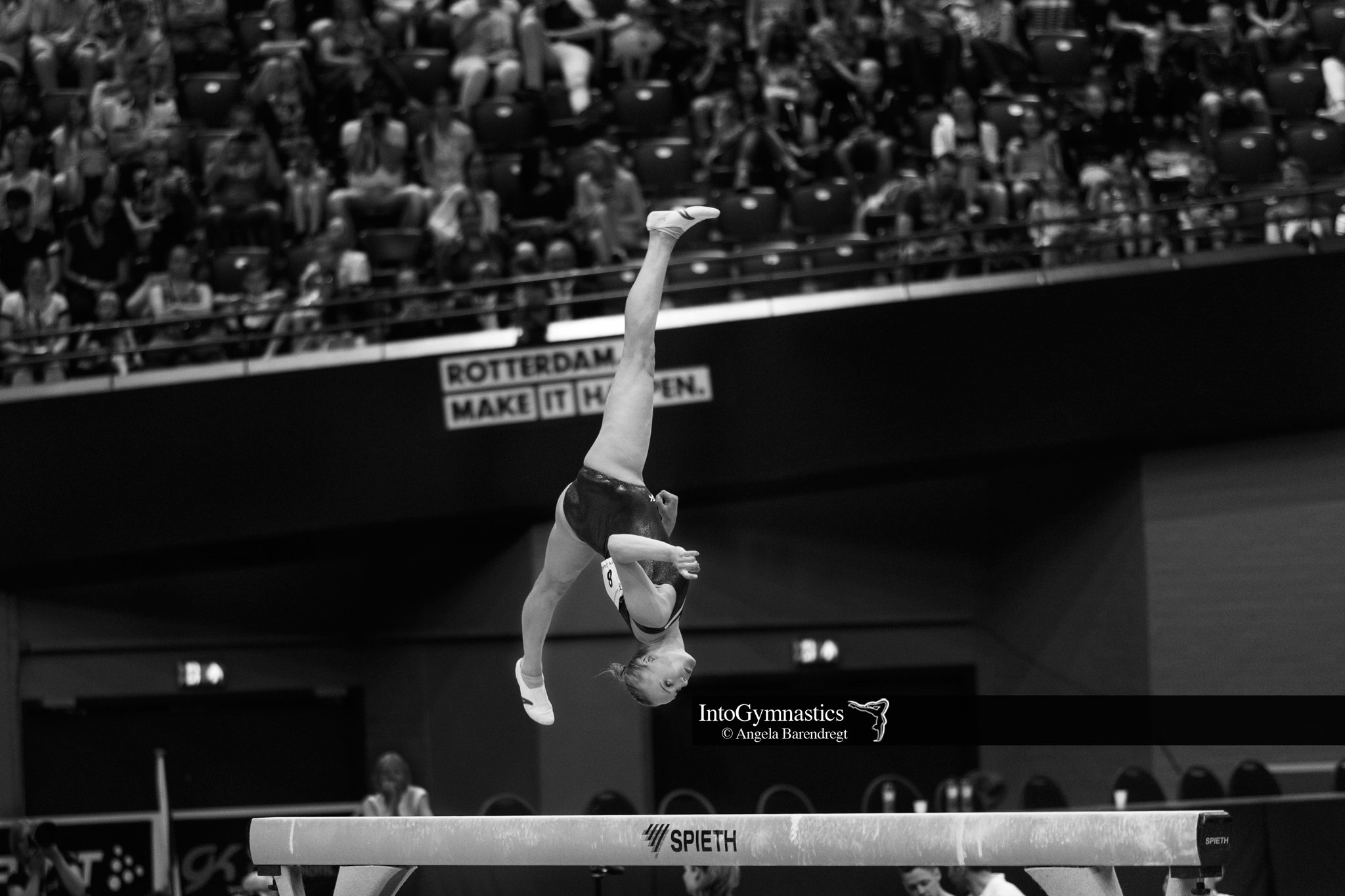 Turnster voert een behendigheidsoefening uit op de evenwichtsbalk in een sporthal met publiek. "Rotterdam Make it Happen" is zichtbaar op de achtergrond.