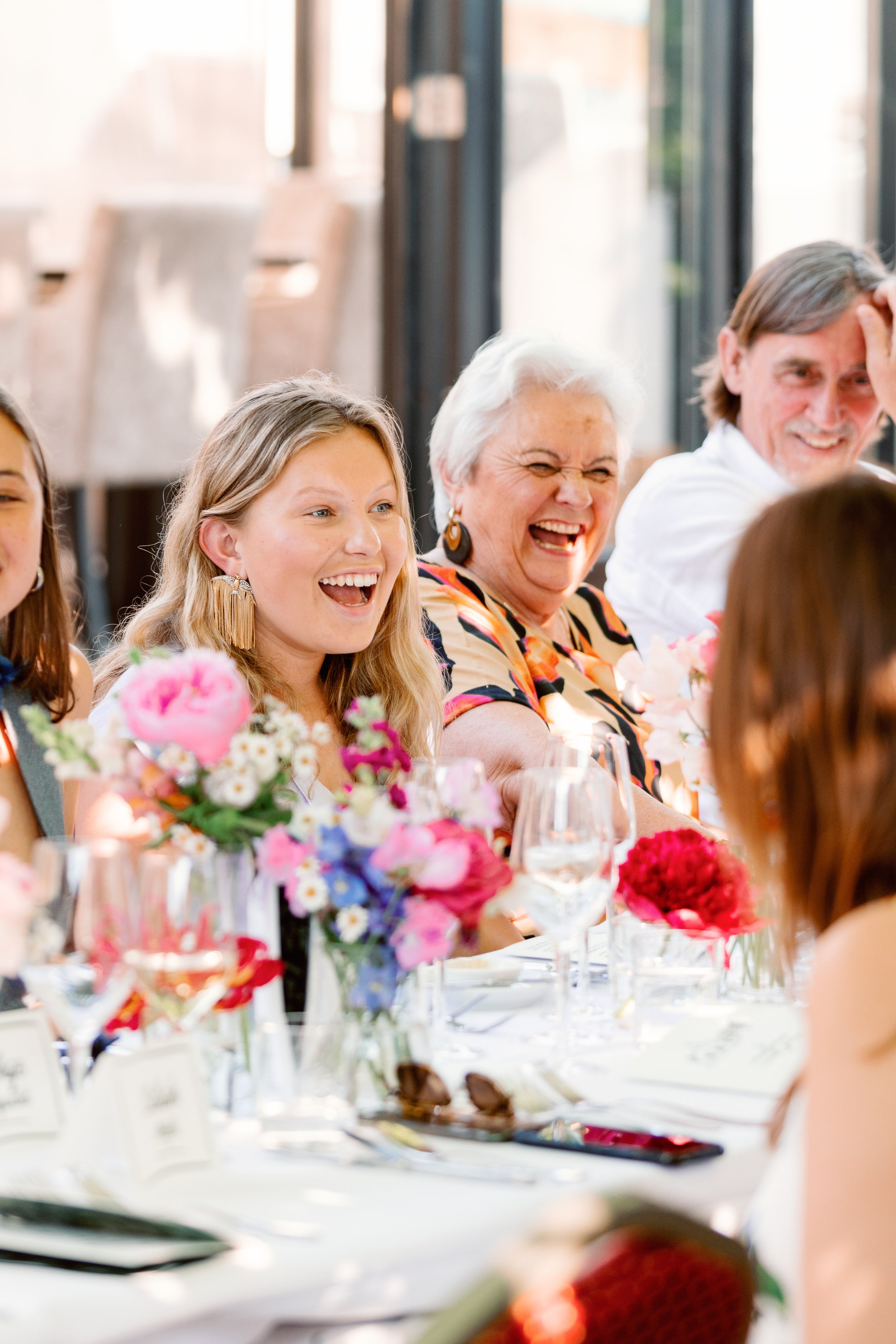 Groep mensen die aan een feestelijk gedekte eettafel zitten en lachen, omringd door kleurrijke bloemen en glazen.