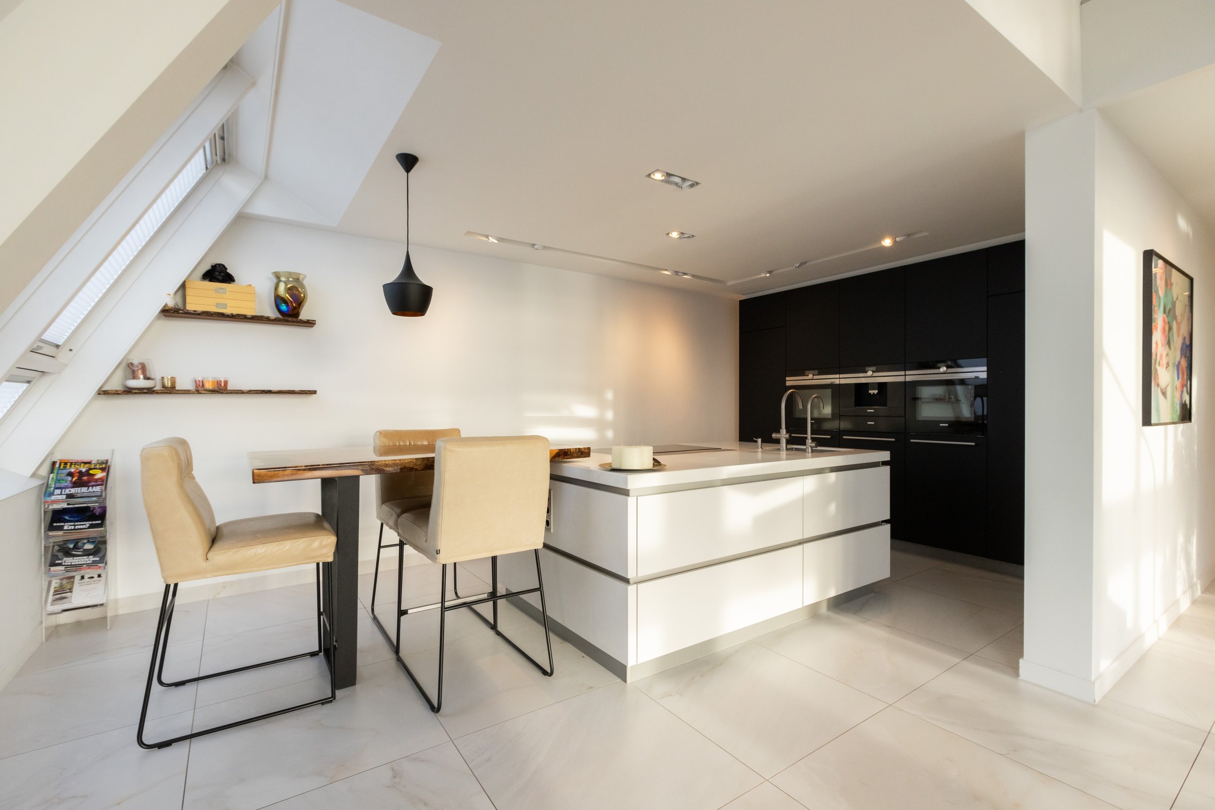 Moderne keuken met eetgedeelte, wit keukeneiland, beige stoelen, hanglamp, open schappen met decoratie, schuine ramen en zwart ingebouwde apparaten.