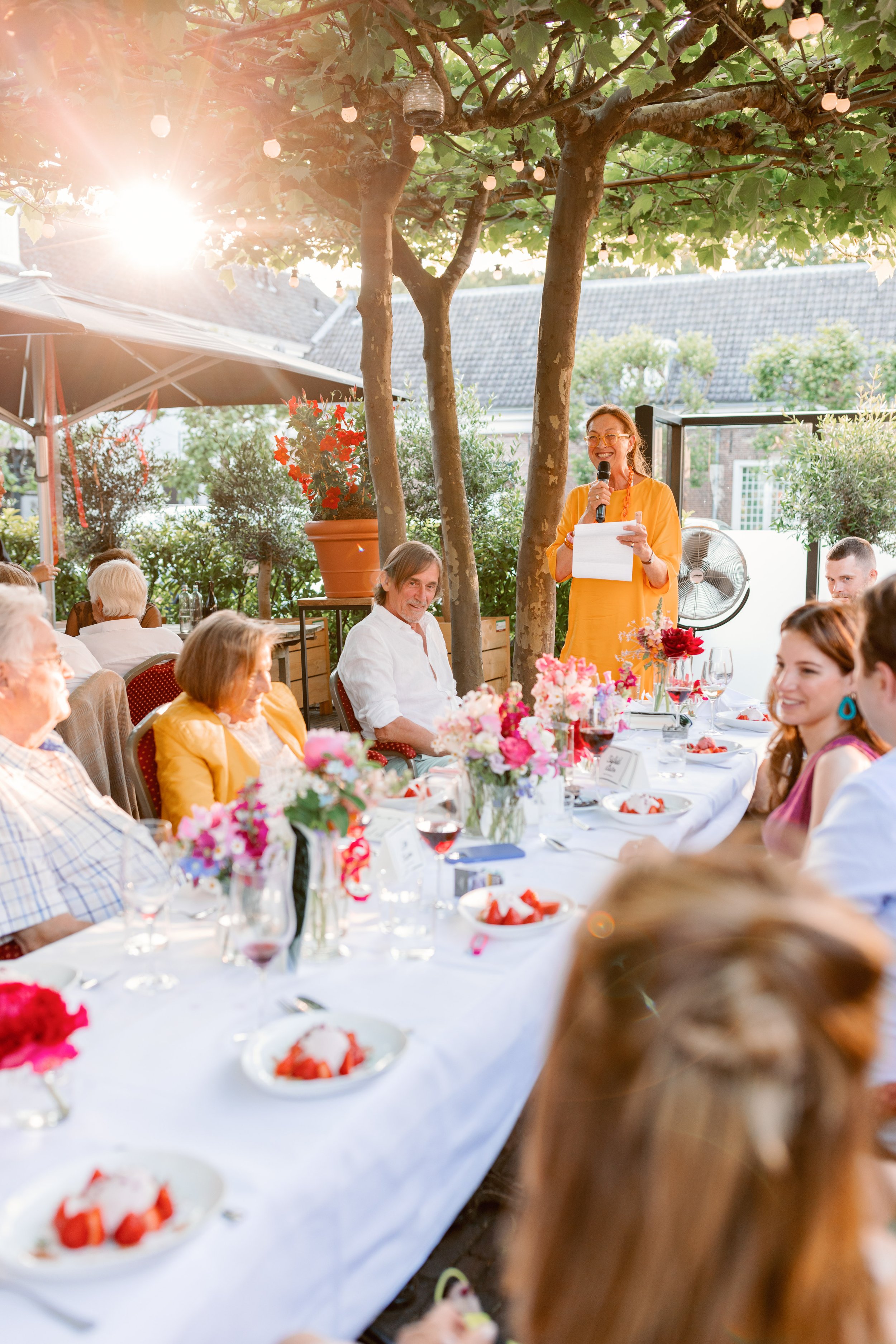 Mensen aan een buitenfeest, vrouw met microfoon houdt toespraak bij versierde tafel, zonsondergang op achtergrond.