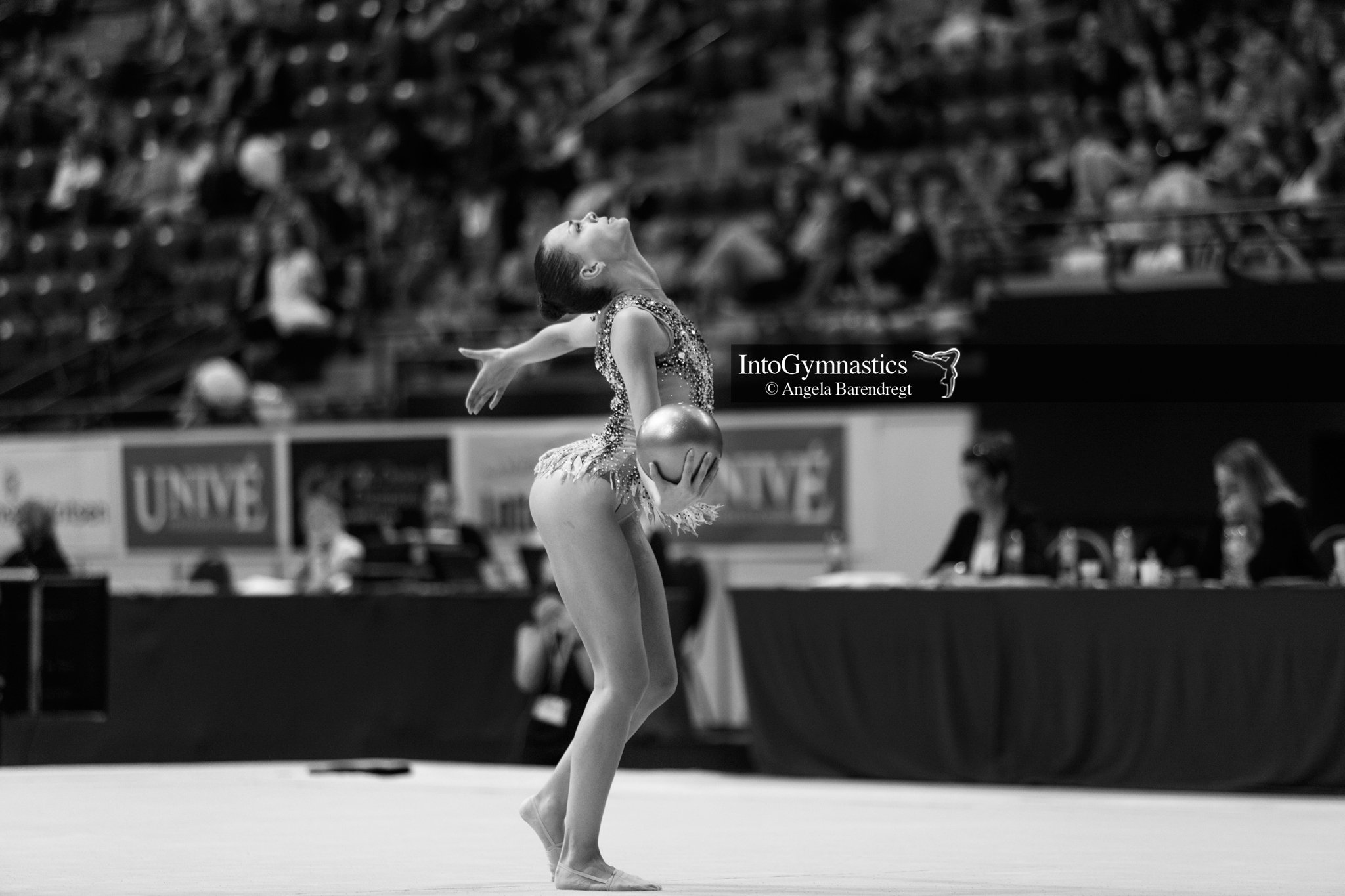 Rhythmische gymnaste voert een oefening uit met een bal tijdens een competitie, met publiek op de achtergrond.
