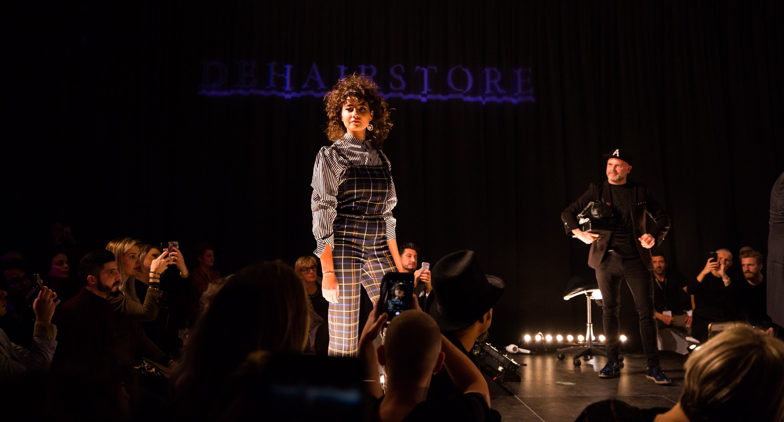 Model loopt op een catwalk tijdens een modeshow, omgeven door een fotograferend publiek. Het model draagt een geruite outfit en heeft krullend haar.