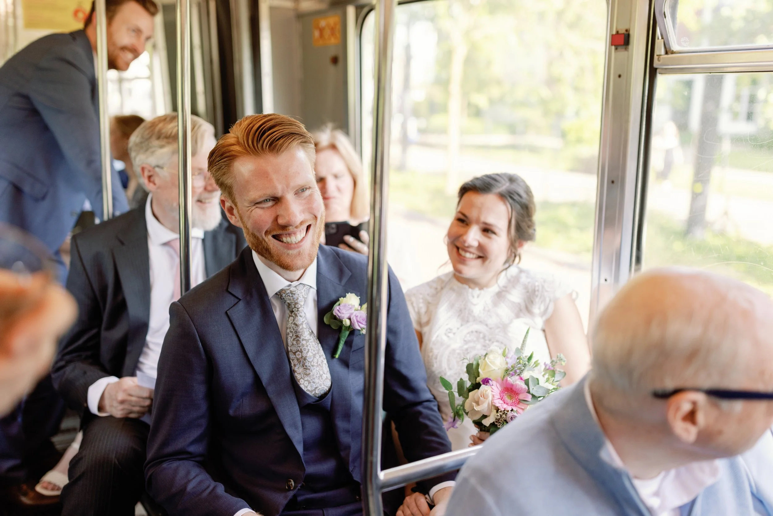 Een bruidspaar zit lachend in een tram, omringd door andere mensen die formeel gekleed zijn. De bruid houdt een boeket met bloemen vast. Ze lijken gelukkig en ontspannen tijdens hun trouwdag.