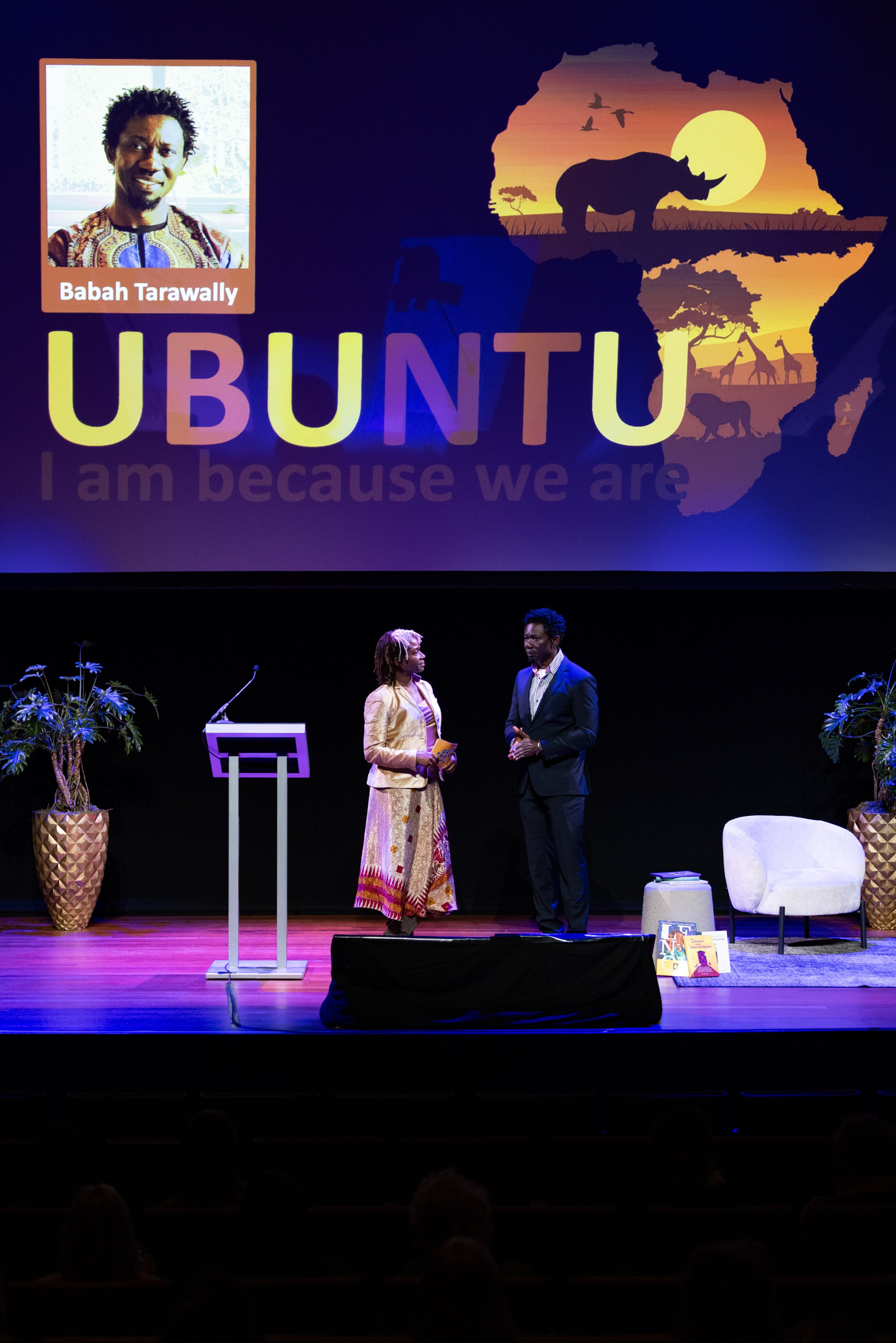 Twee mensen staan op een podium met een grote achtergrondprojectie waarop 'Ubuntu' staat en een afbeelding van Afrika met dieren en een zonsondergang. Er is ook een plant en een stoel op het podium.