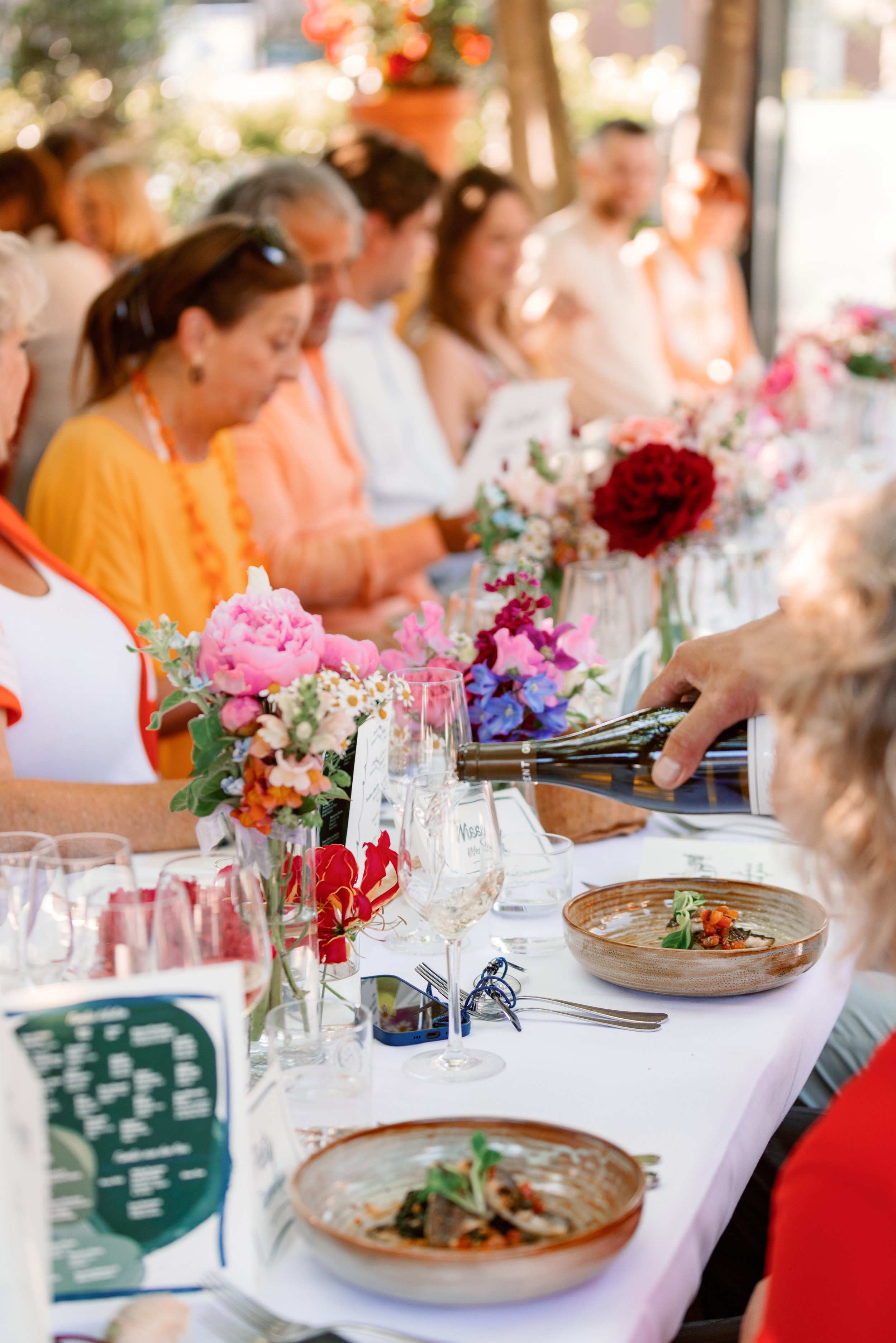 Mensen aan een lange tafel tijdens een feestelijke maaltijd, met bloemen, borden en glazen wijn op tafel.