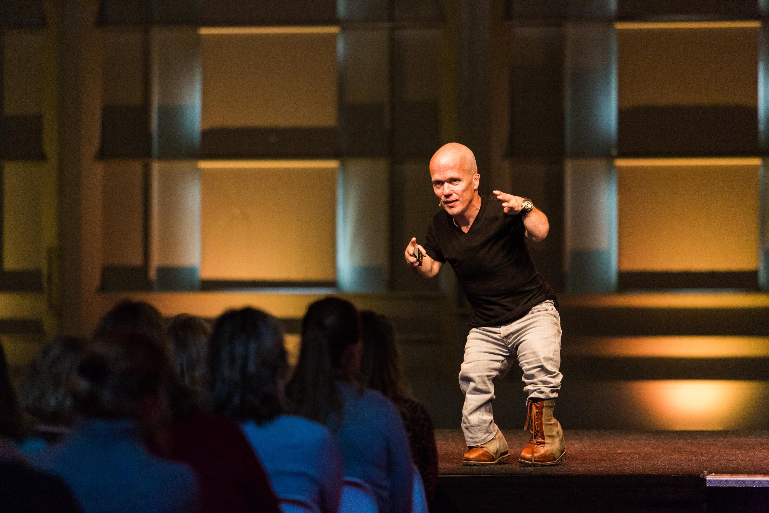 Een man in een zwarte trui en jeans spreekt op een podium voor een zittend publiek. Hij maakt gebaren met zijn handen. De achtergrond is verlicht met warme kleuren.