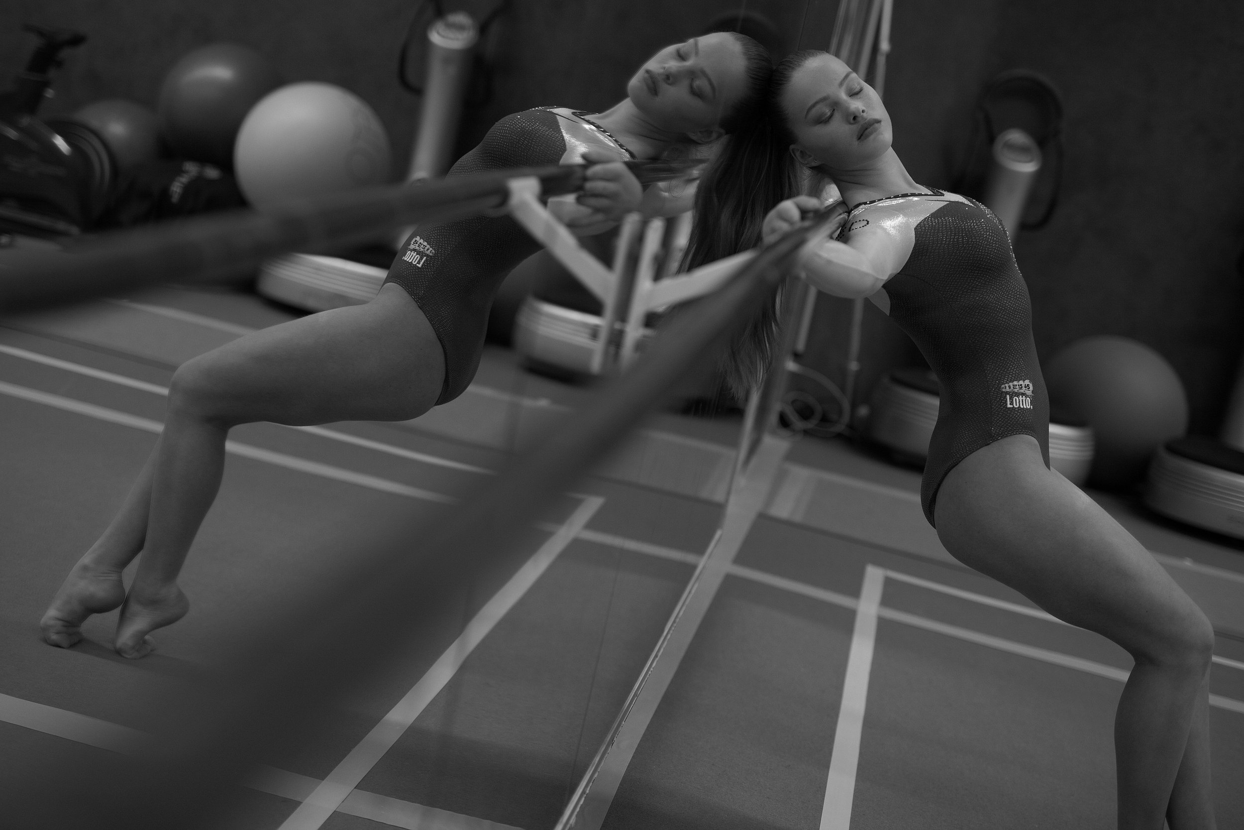 Turnster in zwarte gymnastiekpak maakt een beweging bij een spiegel in een sportzaal met fitnessapparatuur op de achtergrond.