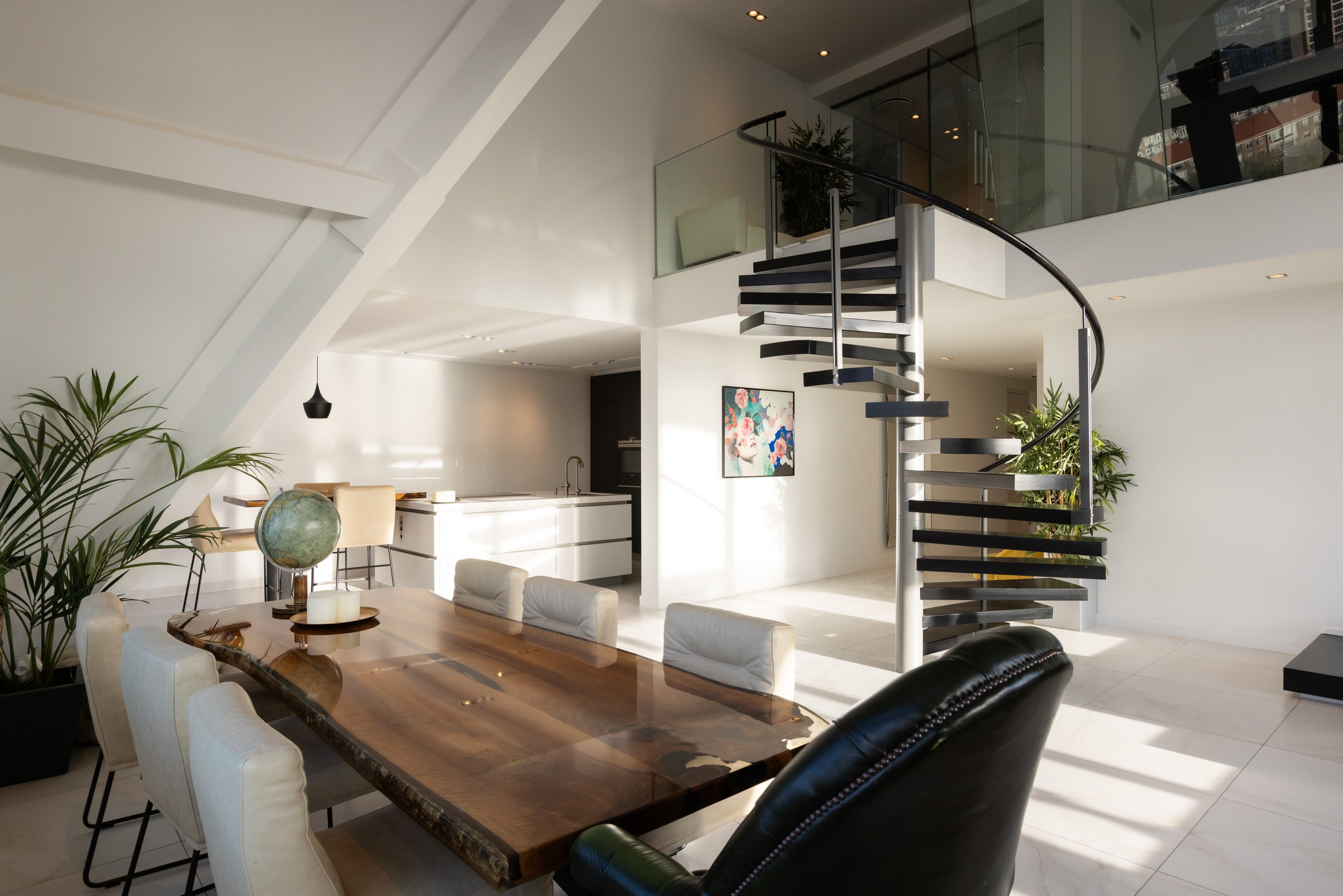 Moderne woonkamer met open keuken, eettafel, glazen trap, en planten.