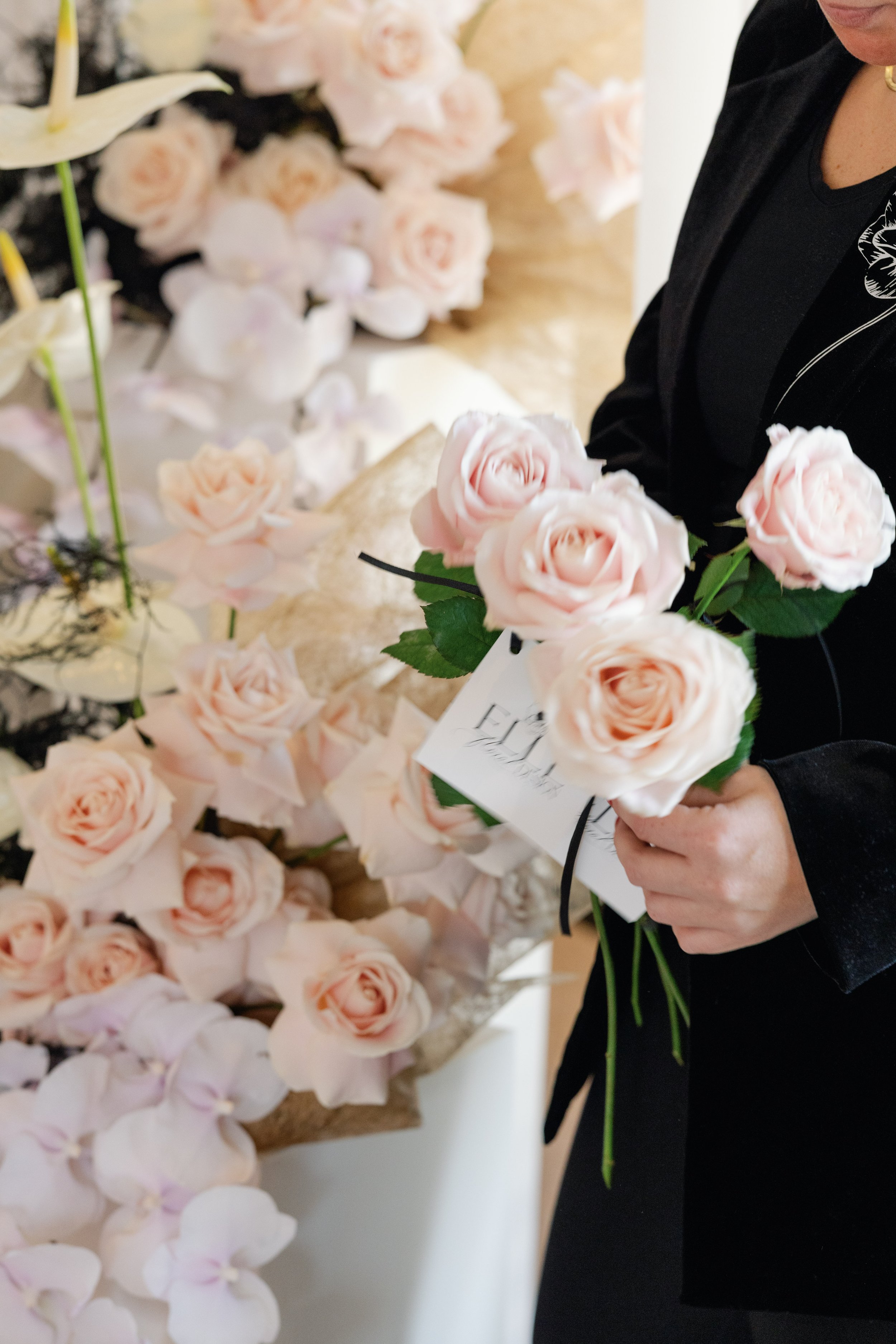 Persoon in zwarte kleding houdt een boeket van lichtroze rozen vast, met meer bloemen op de achtergrond.