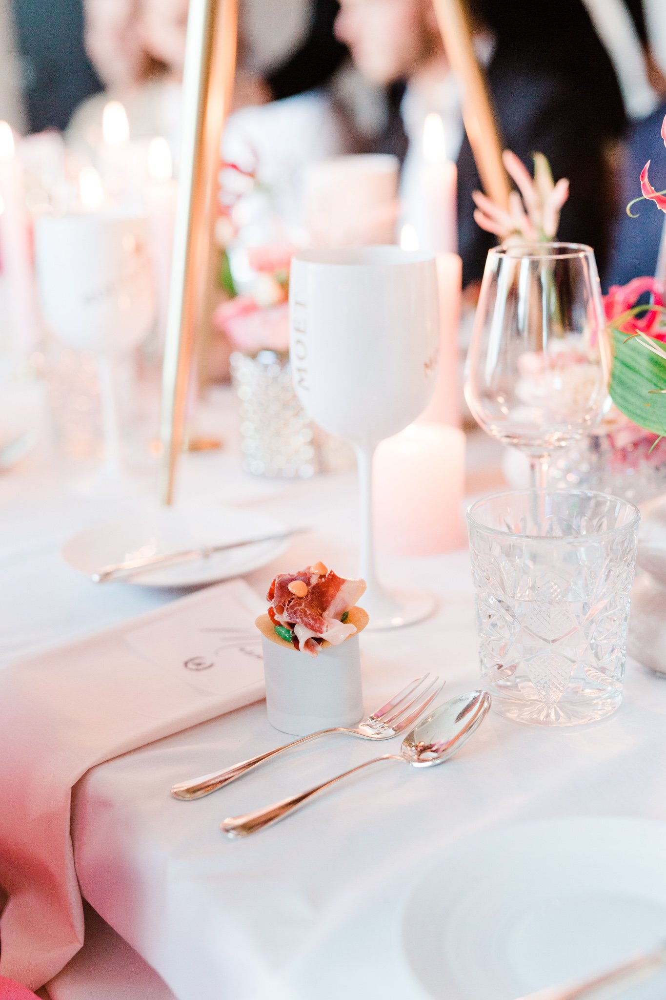 Luxe tafelsetting met witte tafelloper, glanzend bestek, kristallen drinkglas, wijn- en waterglazen en een kleine amuse op een keramische houder. Kaarsen en bloemen sieren de achtergrond.