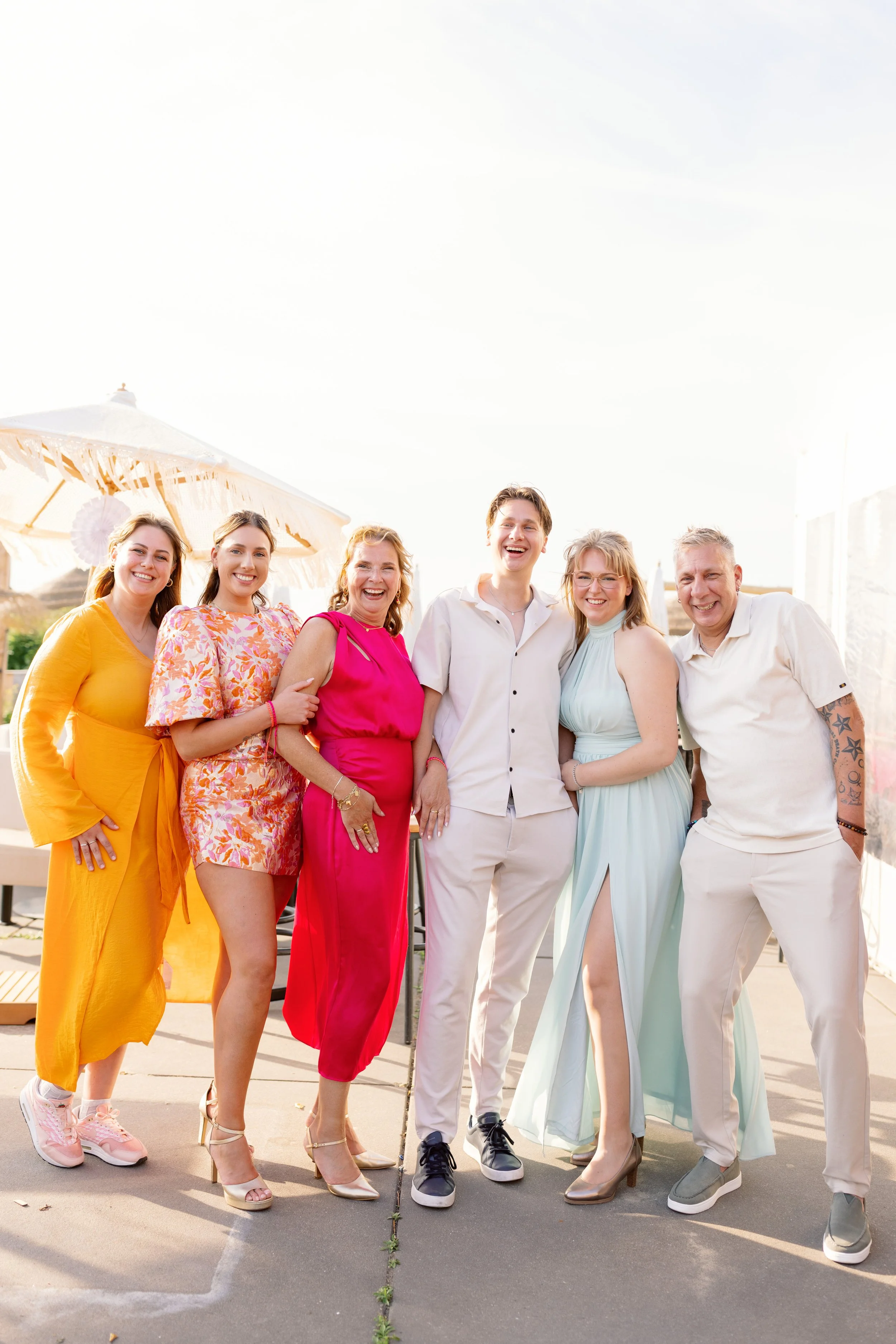 Vrouw in gele jurk, vrouw in bloemenjurk, vrouw in roze jurk, man in wit overhemd, vrouw in lichtblauwe jurk en man in wit overhemd en beige broek staan samen buiten, lachen, onder een parasol met decoraties, zonnige dag.