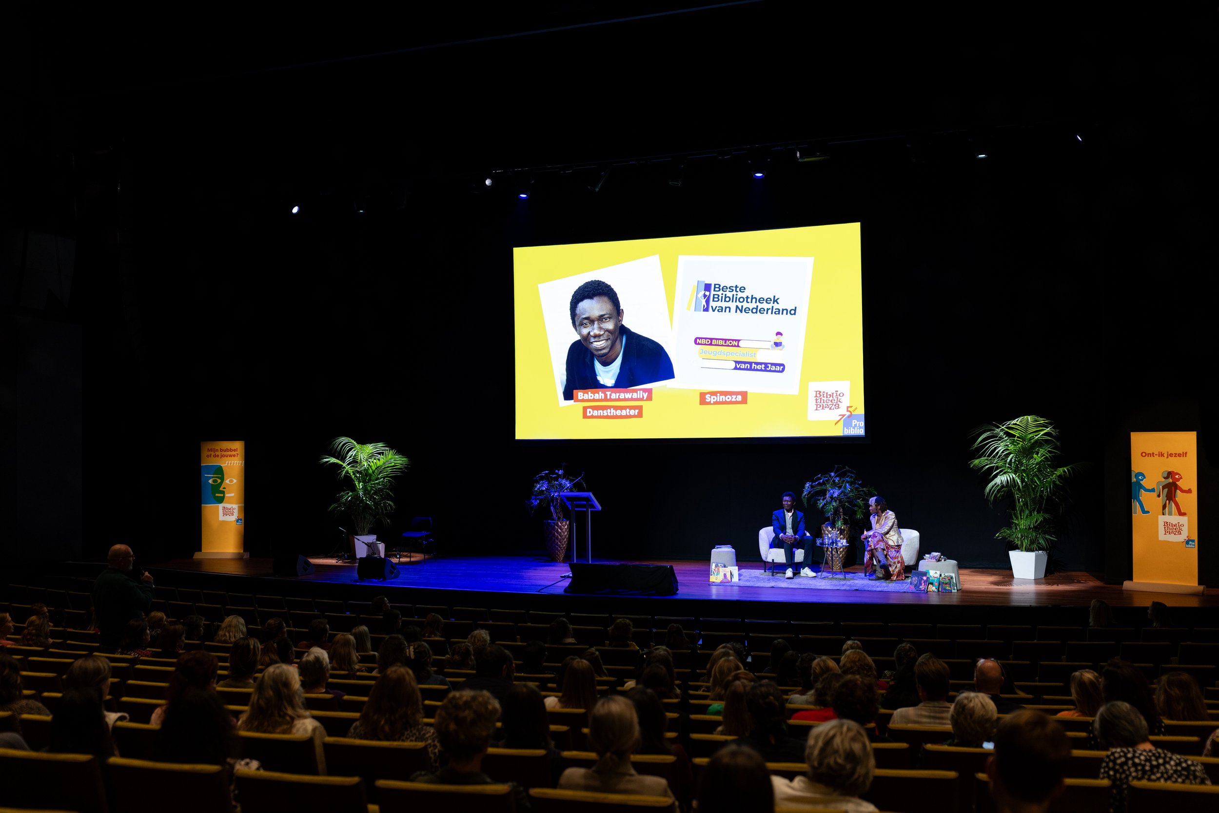Presentatie met publiek in theaterzaal, spreker op podium, groot scherm met tekst 'Beste Bibliotheek van Nederland'. Verschillende mensen zitten in het publiek.