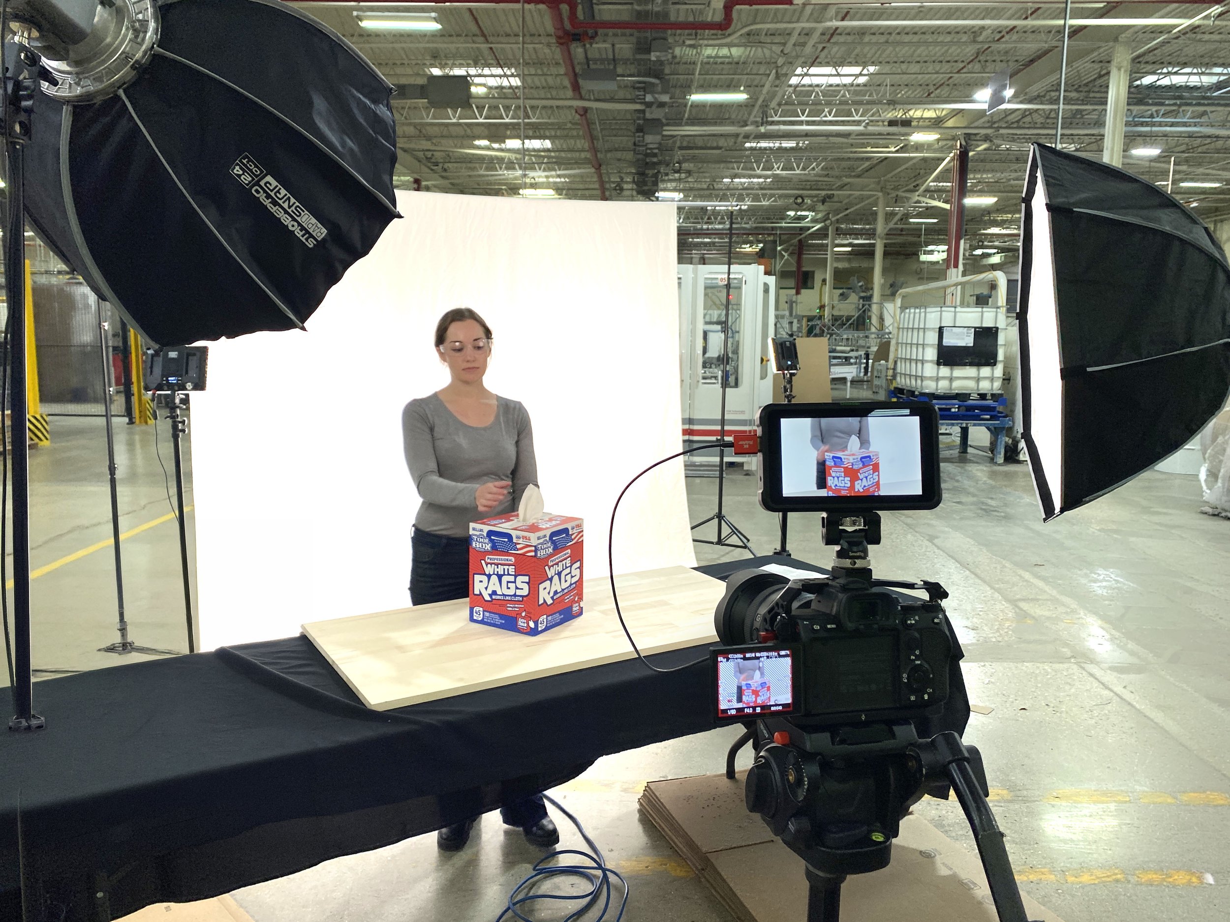 A woman standing behind a box of white rags in a photo studio setup inside a warehouse, with studio lights and equipment around.