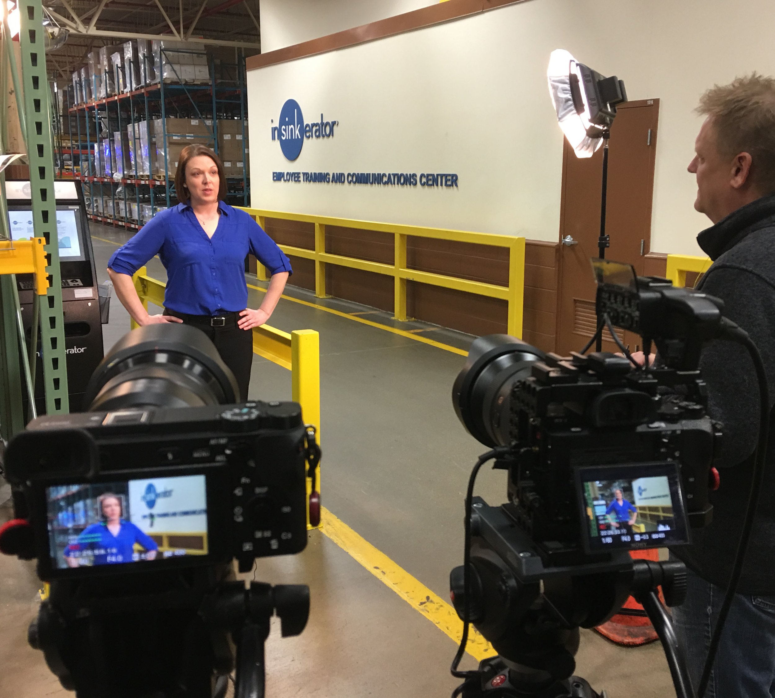A woman in a blue shirt being interviewed by a film crew in front of a sign that says "InSinkErator Employee Training and Communications Center" inside a warehouse.