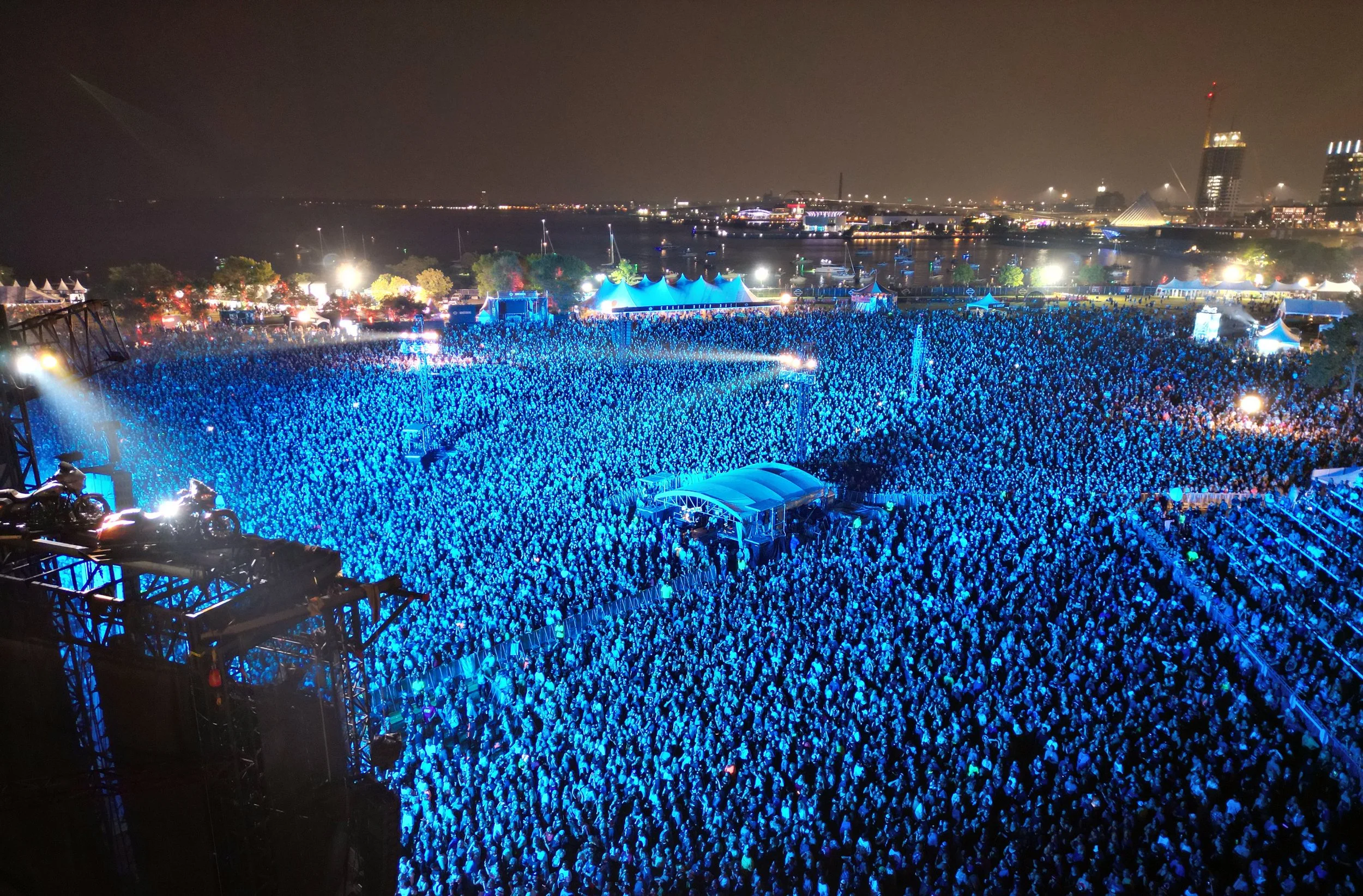 A Harley Davidson Homecoming outdoor concert at night with a crowd of over 70,000 people illuminated by blue lights, set near Lake Michigan with Milwaukee buildings and skyline in the background.