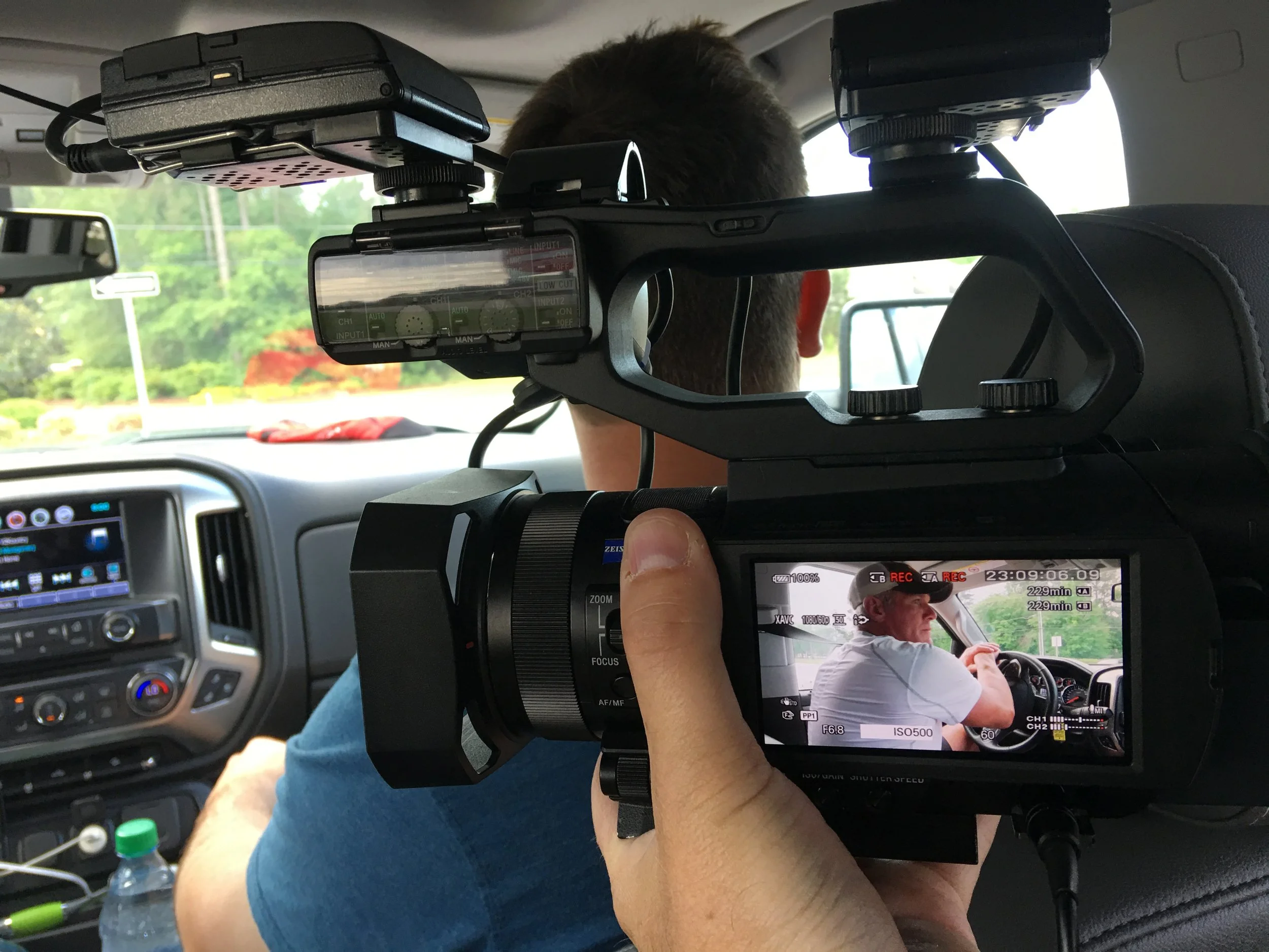 A person operating a professional video camera inside a car, capturing Superbowl MVP Brett Favre driving. The camera is mounted on a stabilizer and the shot is focused on the driver.