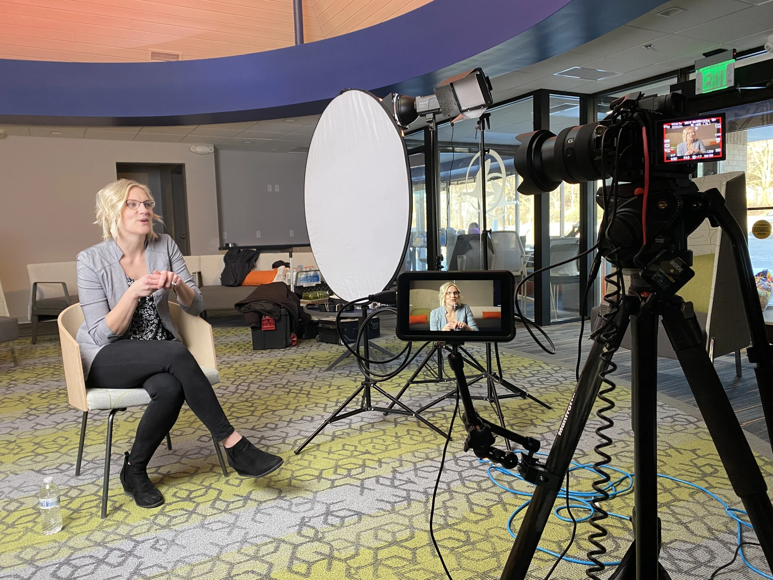 A woman with blonde hair, glasses, and a gray blazer sitting in a chair during a video shoot, with camera equipment, lights, and a white reflector surrounding her in an indoor location with large windows and modern decor.