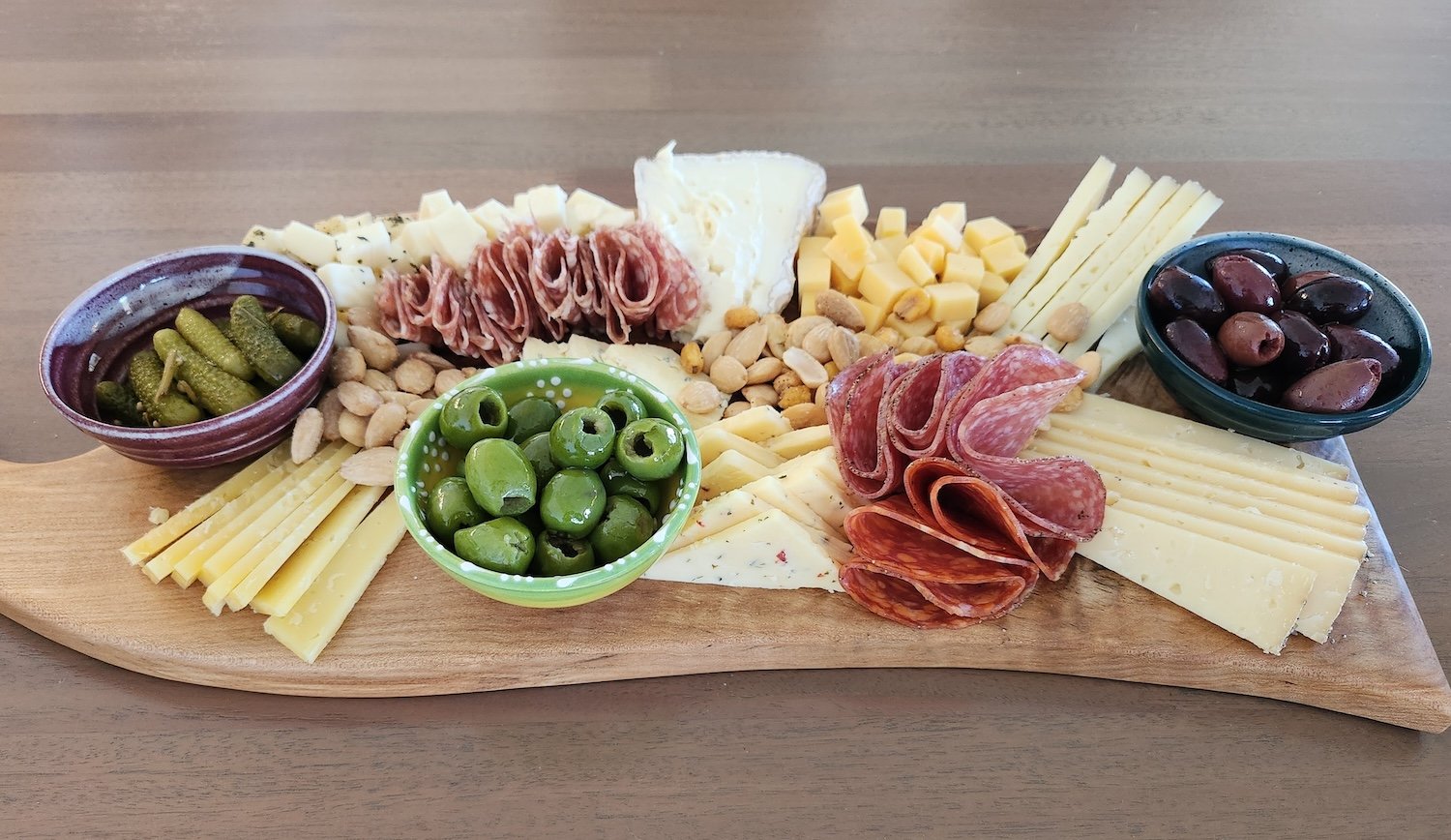Cheese and charcuterie board with various cheeses, cured meats, olives, pickles, nuts, and crackers on a wooden serving board.