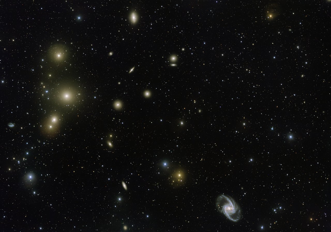 The Fornax Galaxy Cluster is one of the closest of such groupings beyond our Local Group of galaxies. This new VLT Survey Telescope image shows the central part of the cluster in great detail. At the lower-right is the elegant barred-spiral galaxy NGC 1365 and to the left the big elliptical NGC 1399. – Image Credit: ESO. Acknowledgement: Aniello Grado and Luca Limatola