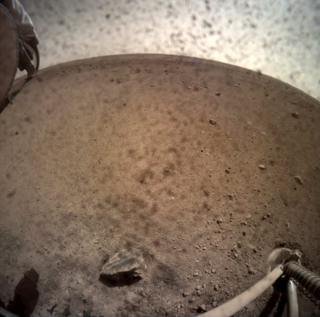 NASA’s InSight spacecraft flipped open the lens cover on its Instrument Context Camera (ICC) on Nov. 30, 2018, and captured this view of Mars. Located below the deck of the InSight lander, the ICC has a fisheye view, creating a curved horizon. Some clumps of dust are still visible on the camera’s lens. One of the spacecraft’s footpads can be seen in the lower right corner. The seismometer’s tether box is in the upper left corner. - Image Credit: NASA/JPL-Caltech