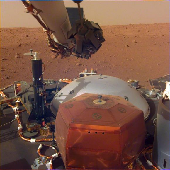 This image was taken by the InSight Lander’s Instrument Deployment Camera mounted on the lander’s robotic arm. The stowed grapple on the end of the arm is folded in, but it will unfold and be used to deploy the lander’s science instrument. The copper-colored hexagonal object is the protective cover for the seismometer, and the grey dome behind it is a wind and thermal shield, which will be placed over the seismometer after its deployed. The black cyliner on the left is the heat probe, which will drill up to 5 meters into the Martian surface. - Image Credit: NASA/JPL-Caltec
