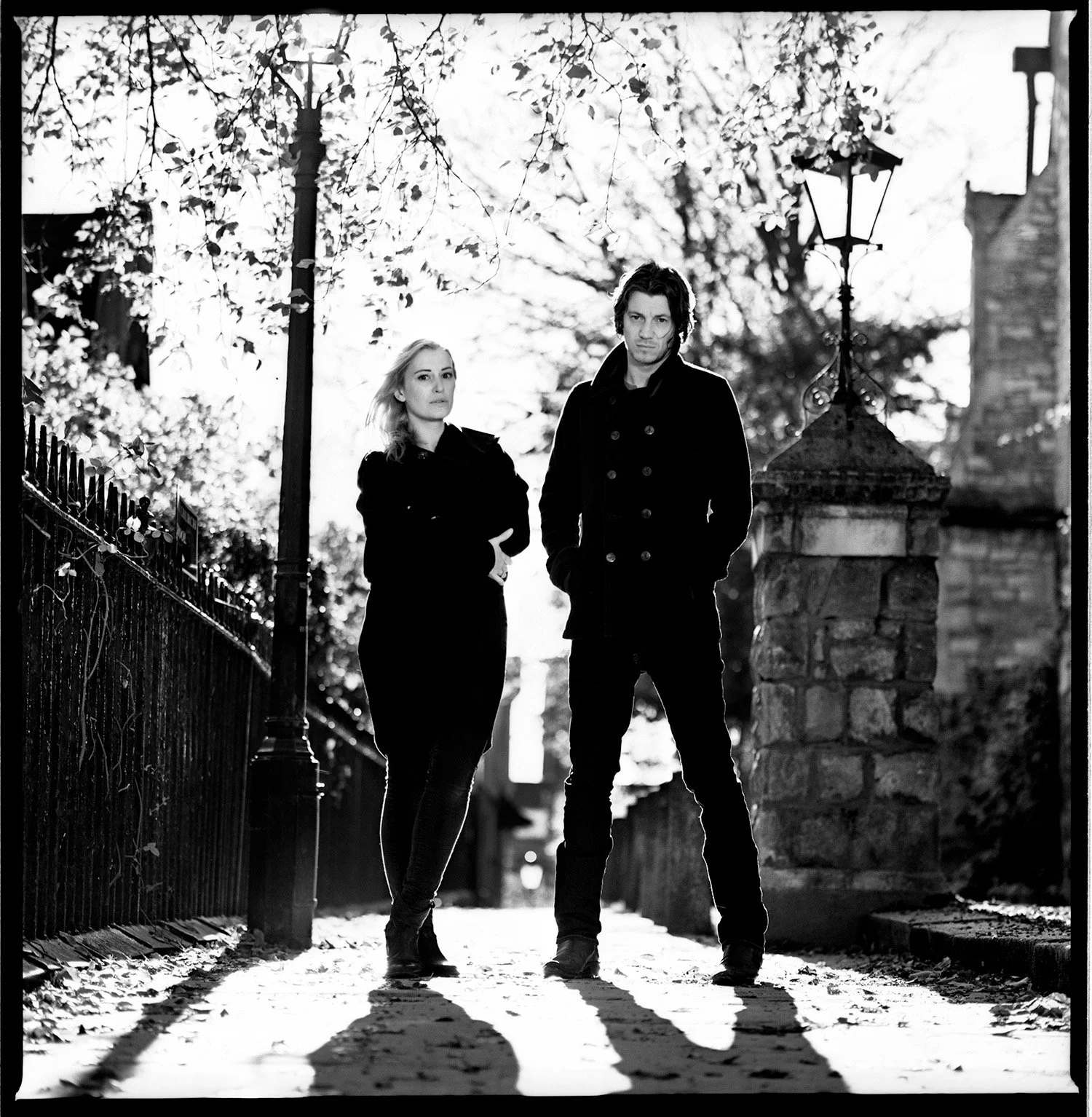 Black-and-white photo of a man and woman standing on a sidewalk, casting long shadows, with trees, street lamp, and stone fences in the background.