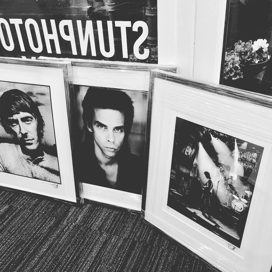 Three framed photographs, two portraits of male musicians and a live concert shot, are displayed on the floor leaning against a wall.
