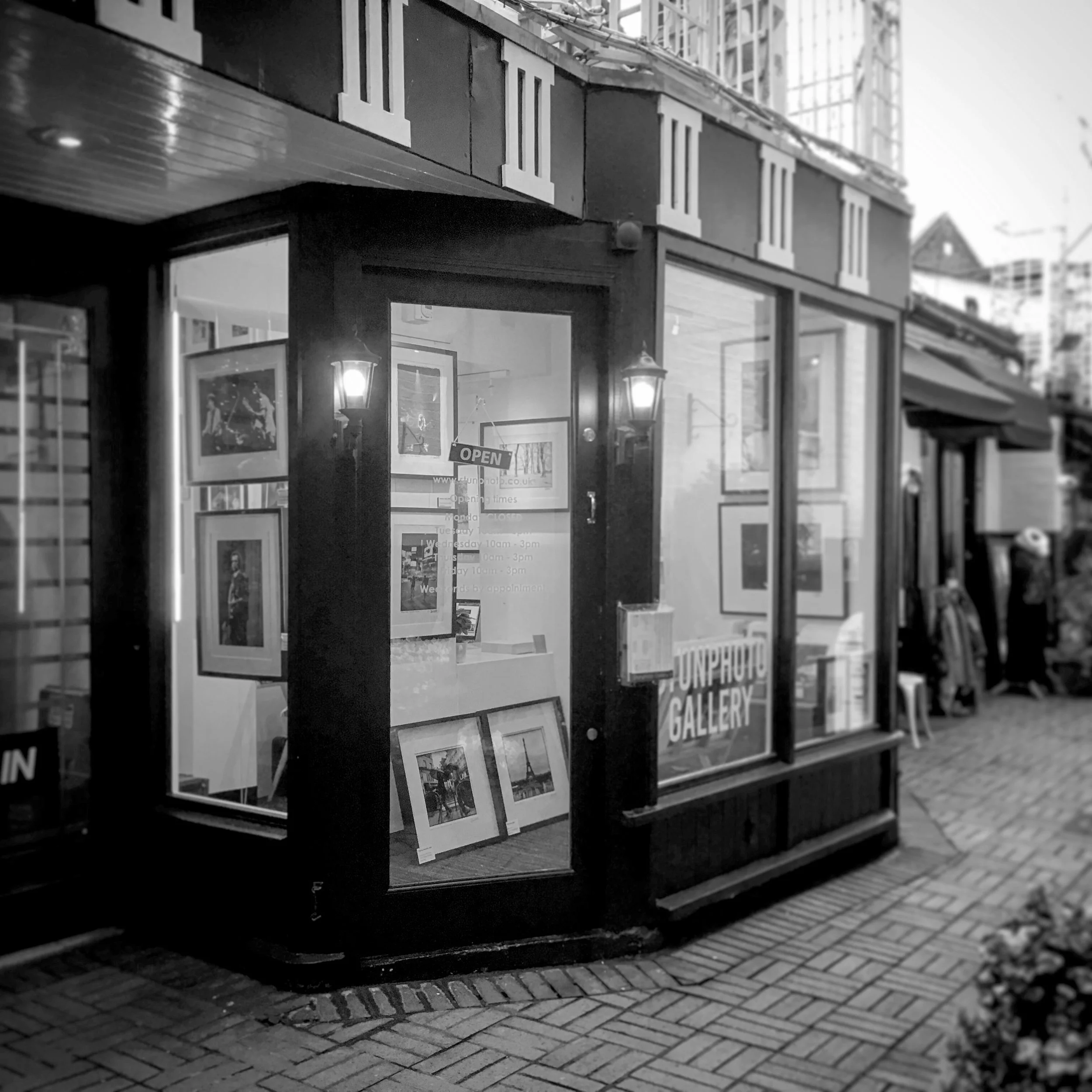 Black and white photo of the exterior of an art gallery with framed artwork on display and an 'OPEN' sign on the door. There are lantern-style lights near the entrance and a brick sidewalk in front.
