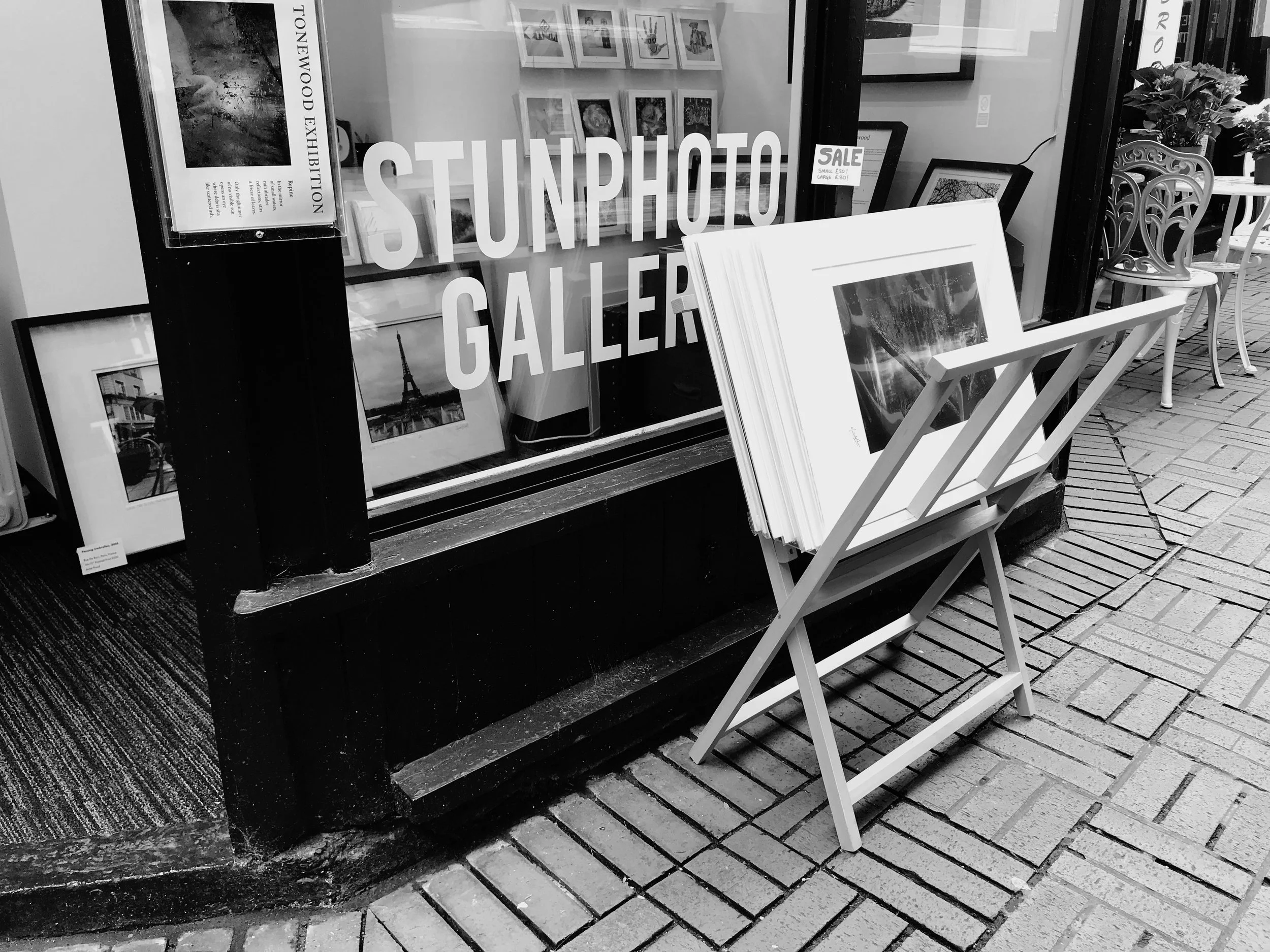 Black and white photo of the storefront of a gallery with large text reading 'Sunhouse Gallery'. There is a display of framed pictures inside and outside the shop, with a sign indicating a sale on artwork. Outside, a white bench holds large art prints, and to the right, there's a decorative outdoor chair placed on a tiled sidewalk.