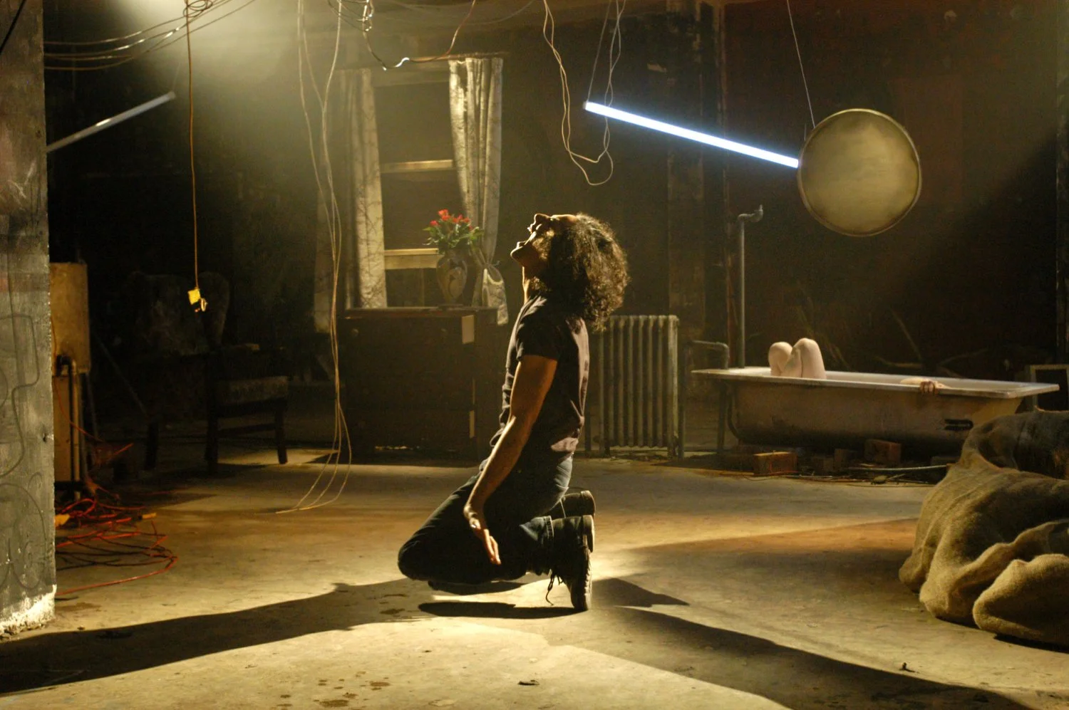 A woman with curly hair kneels on the floor in a dimly lit, industrial-style room, with her head tilted back and eyes closed, appearing to be in a moment of emotional surrender or dance, surrounded by various objects including a bathtub with a small child lying inside, a chair, a flower vase on a table, and hanging wires and lights.