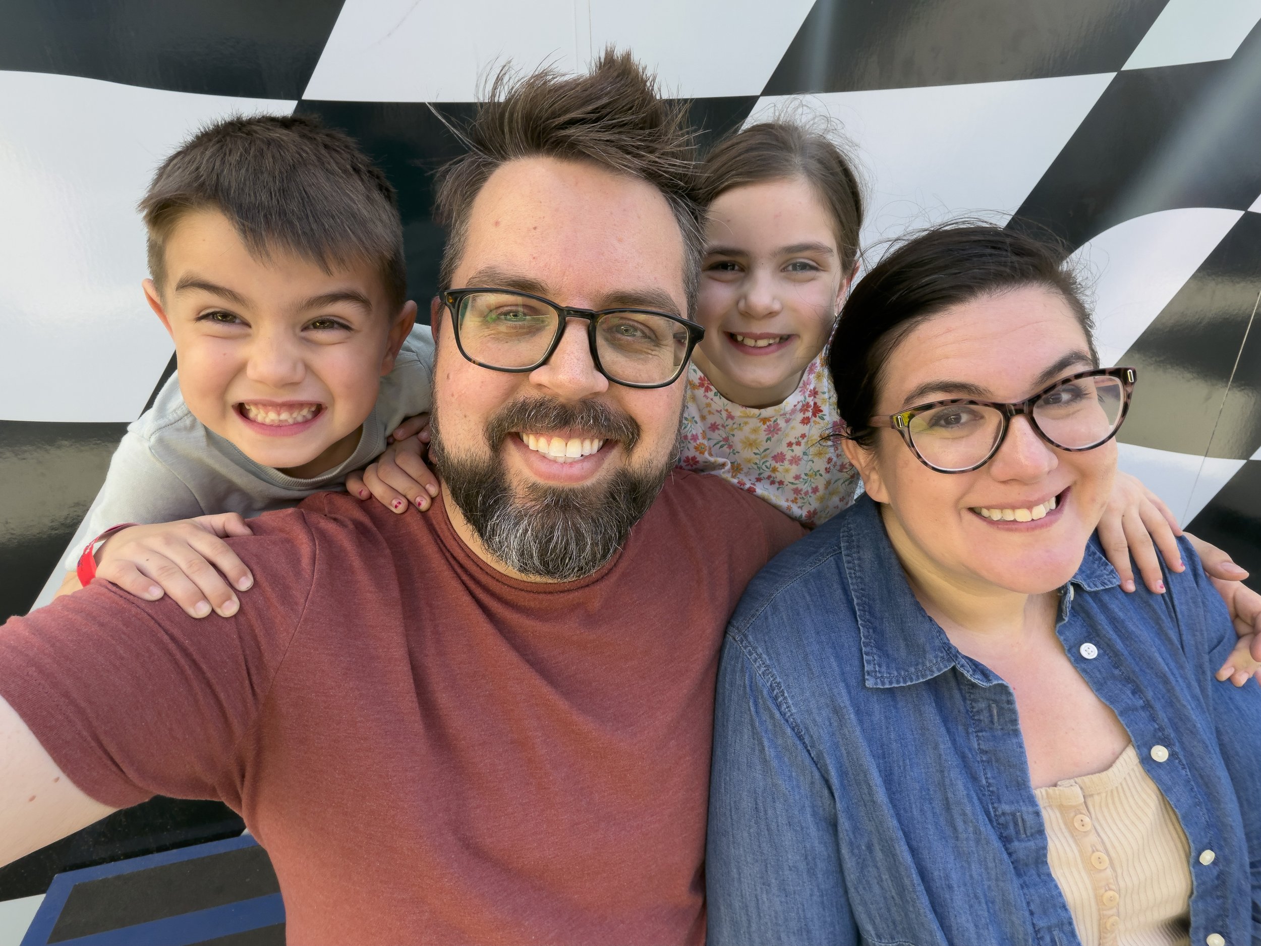A happy family taking a close-up selfie together, including a man with glasses, two women, and two children, in front of a black and white checkered background.