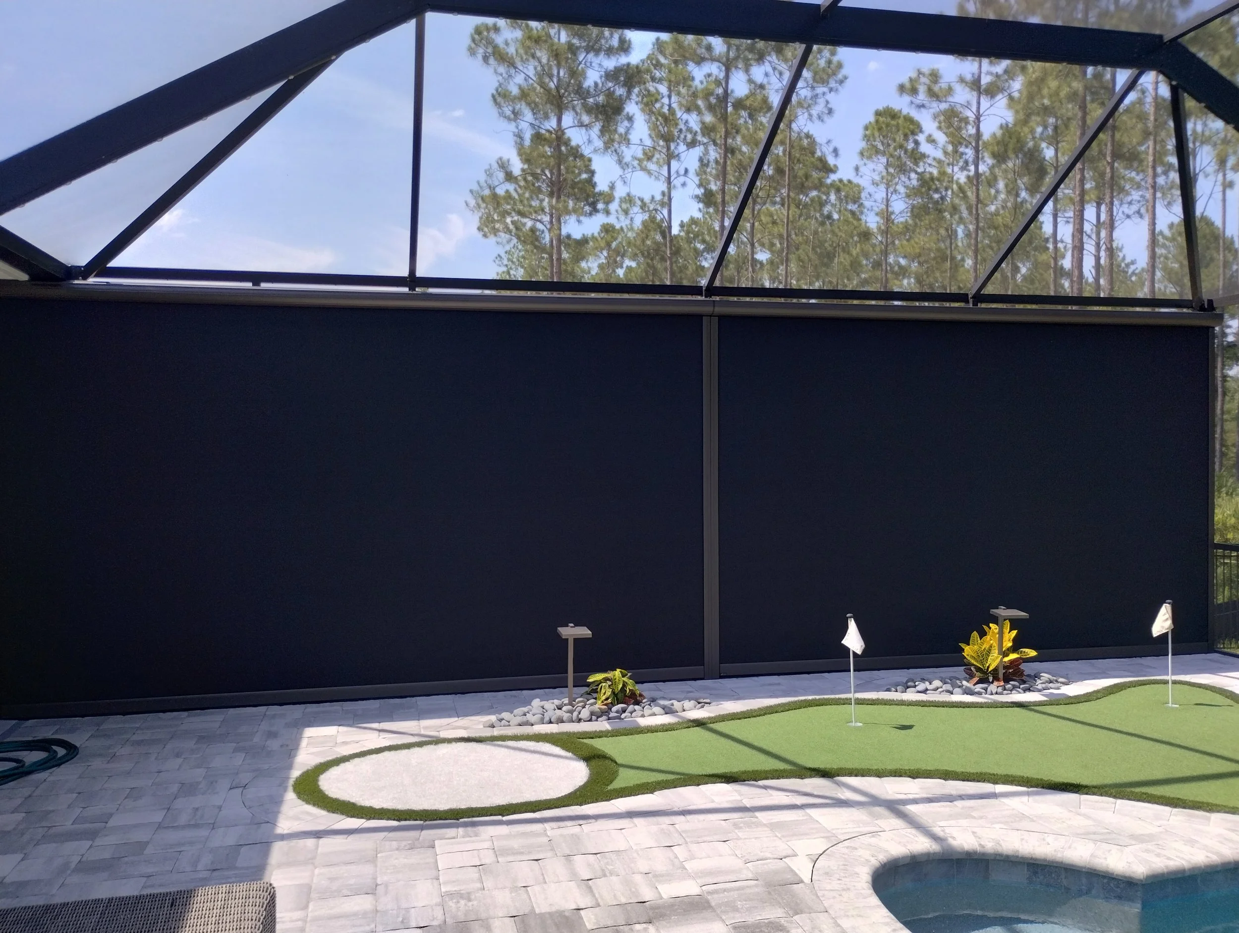 Backyard patio with a putting green, small plants, and a privacy retractable shade wall in a pool screen enclosure.