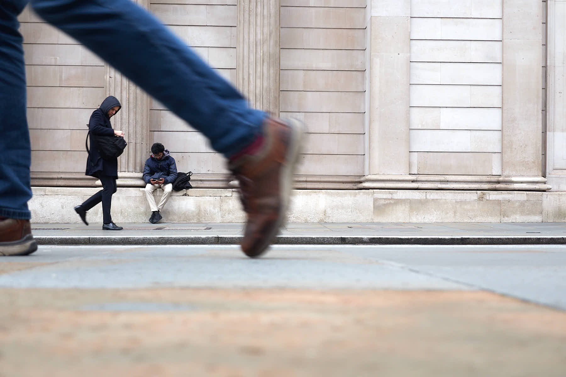 A man is sitting on a low wall in front of the Bank of England in London. He is framed in the legs of a passer by closer to the camera which is at a very low level. There is also a second man framed in the same space