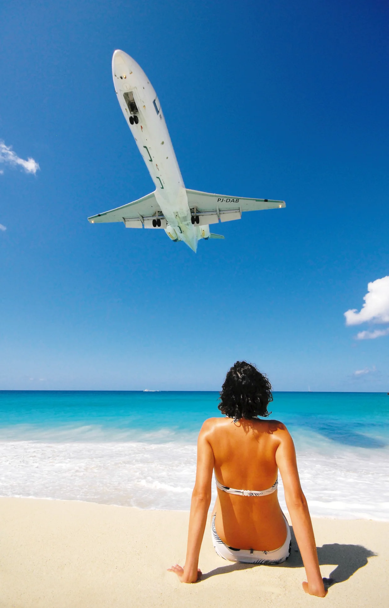 A young woman sitting on a beach, seen from behind while watching a low flying aircraft is very close to the beach
