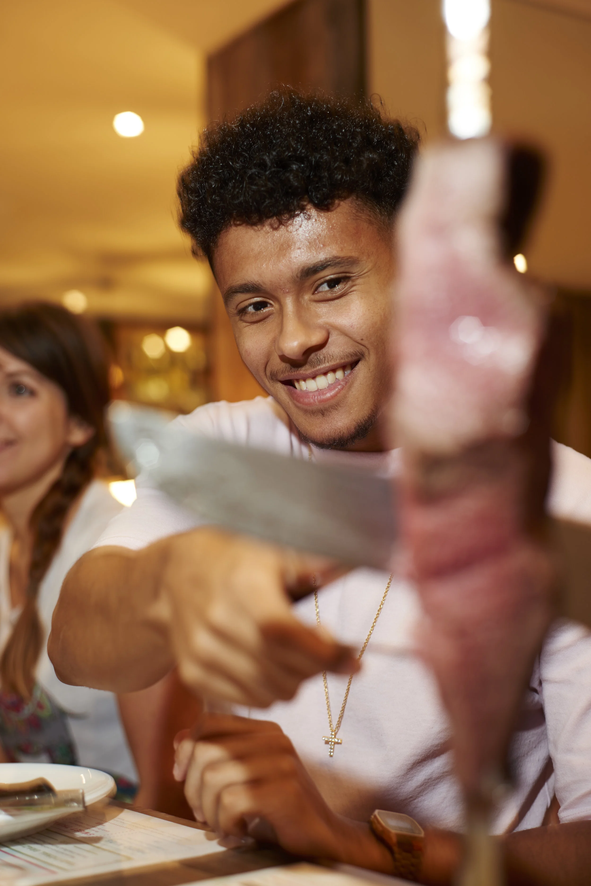 A smiling young man is using tongs to receive a slice of meat carved from a skewer being served at a table in a restaurant