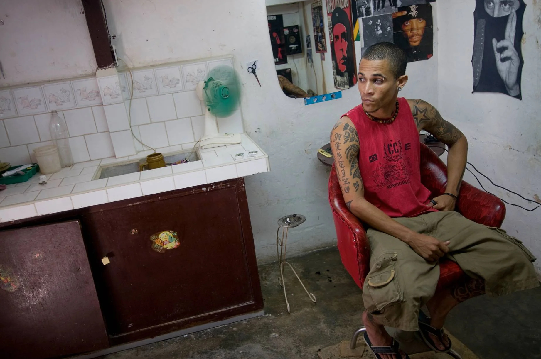 Wide angle portrait of a young man with tattooed arms wearing a red vest top sitting in a barbers chair next to a wash basin in a makeshift barbershop in Havana, Cuba
